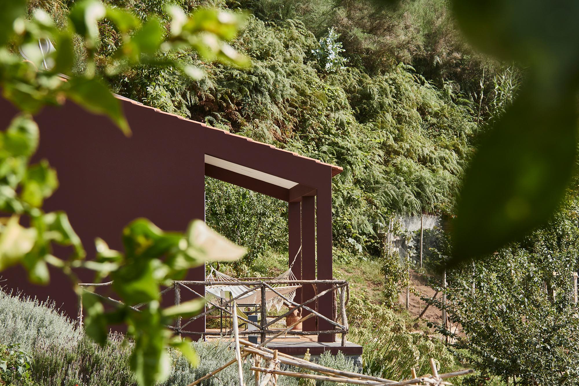 A dark red building with a covered porch and hammocks, seen past out-of-focus leaves in the foreground, set against a wooded hillside.