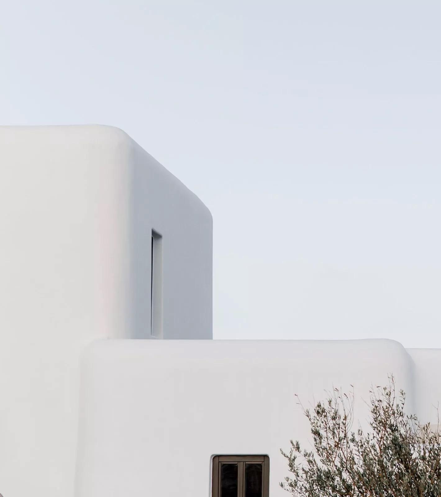 A white cycladic building's curved corner with a narrow dark window, pale sky above and an olive branch crossing the lower right.