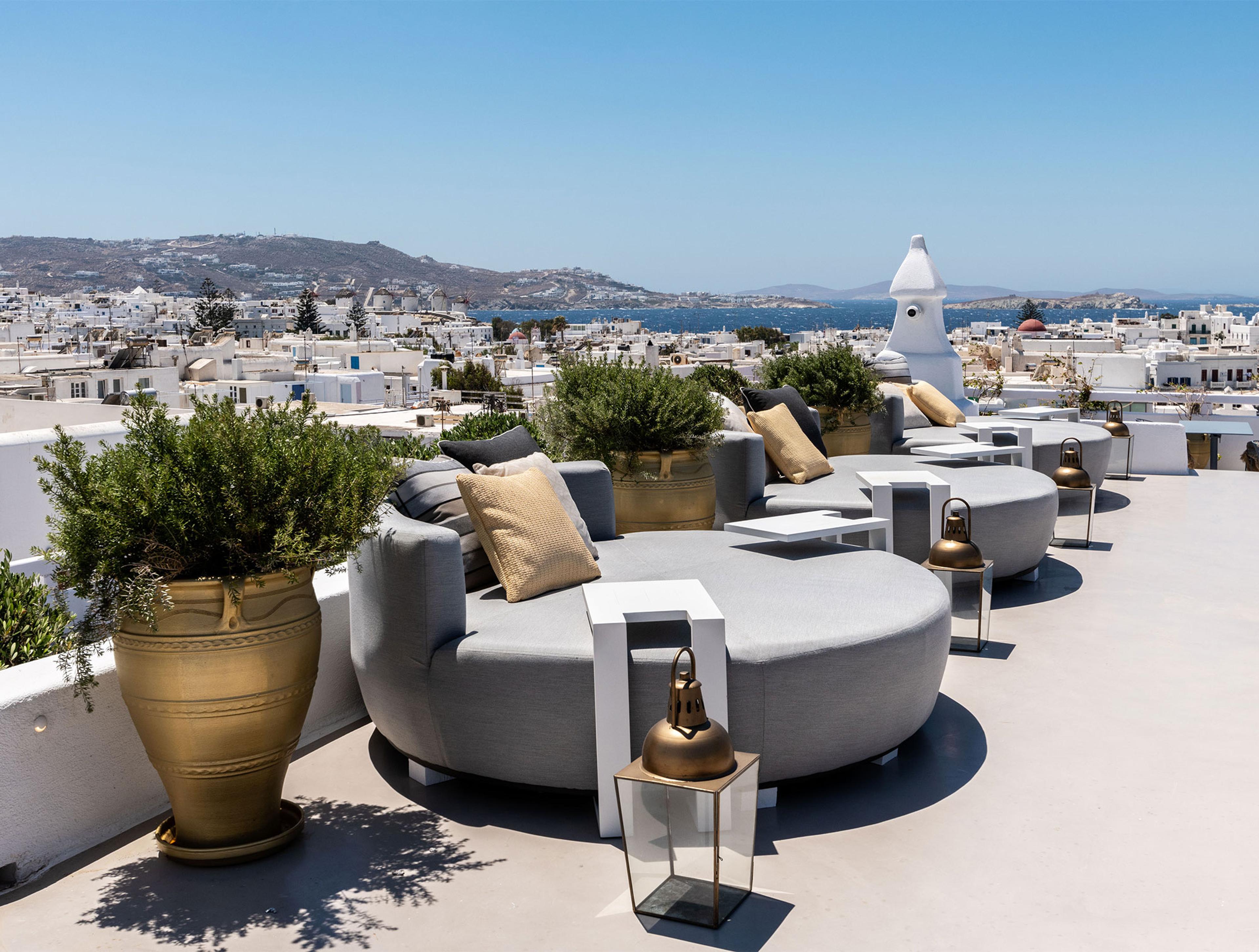 A sunlit rooftop terrace with round sofas, potted plants and brass lanterns, overlooking the white rooftops of Mykonos town and the sea.