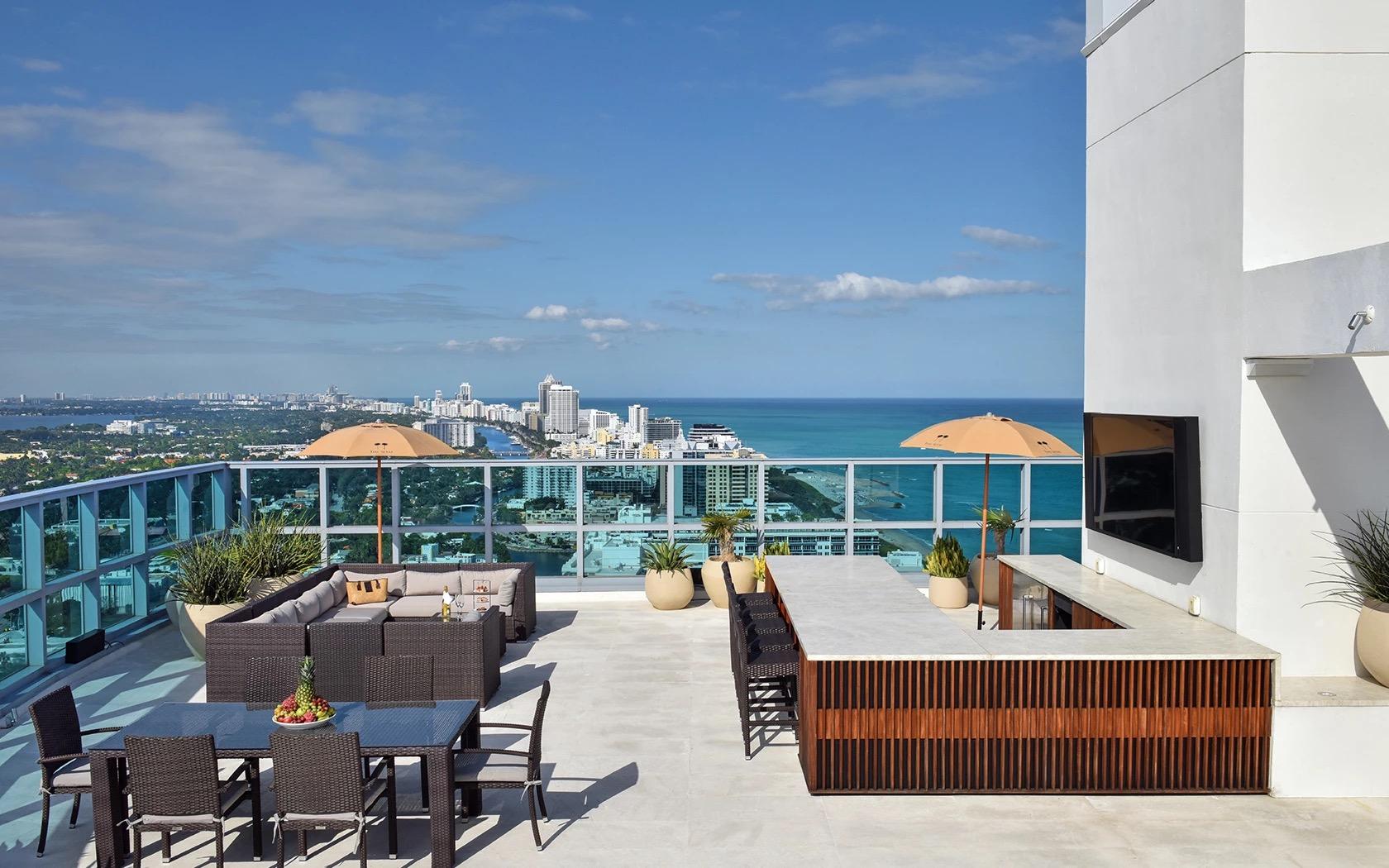 A rooftop terrace with sofas, a dining table and a bar, overlooking the Miami Beach skyline and ocean under a clear sky.