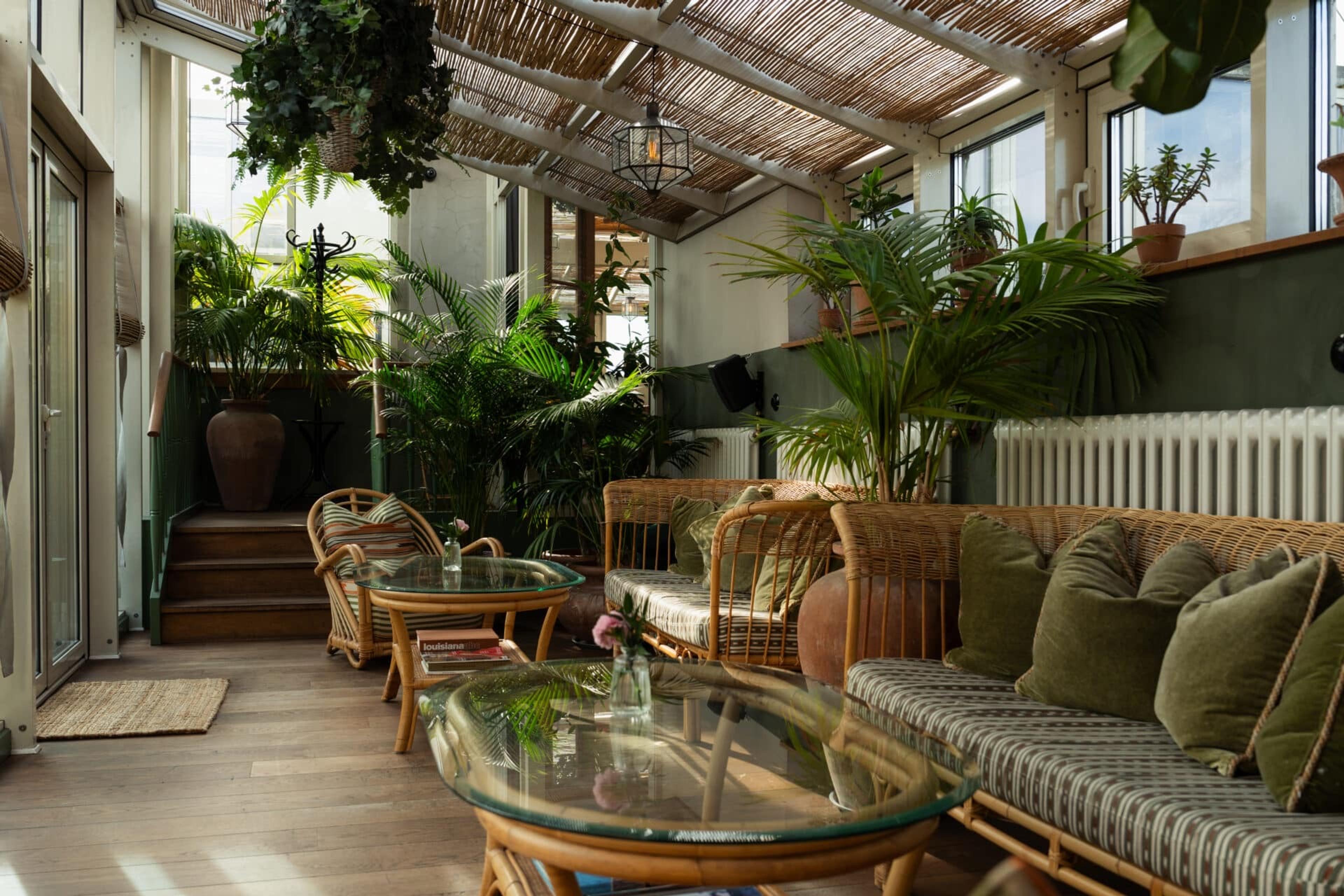 A glass-roofed lounge filled with potted palms and ferns, with rattan sofas and armchairs around glass coffee tables and a staircase at the far end.