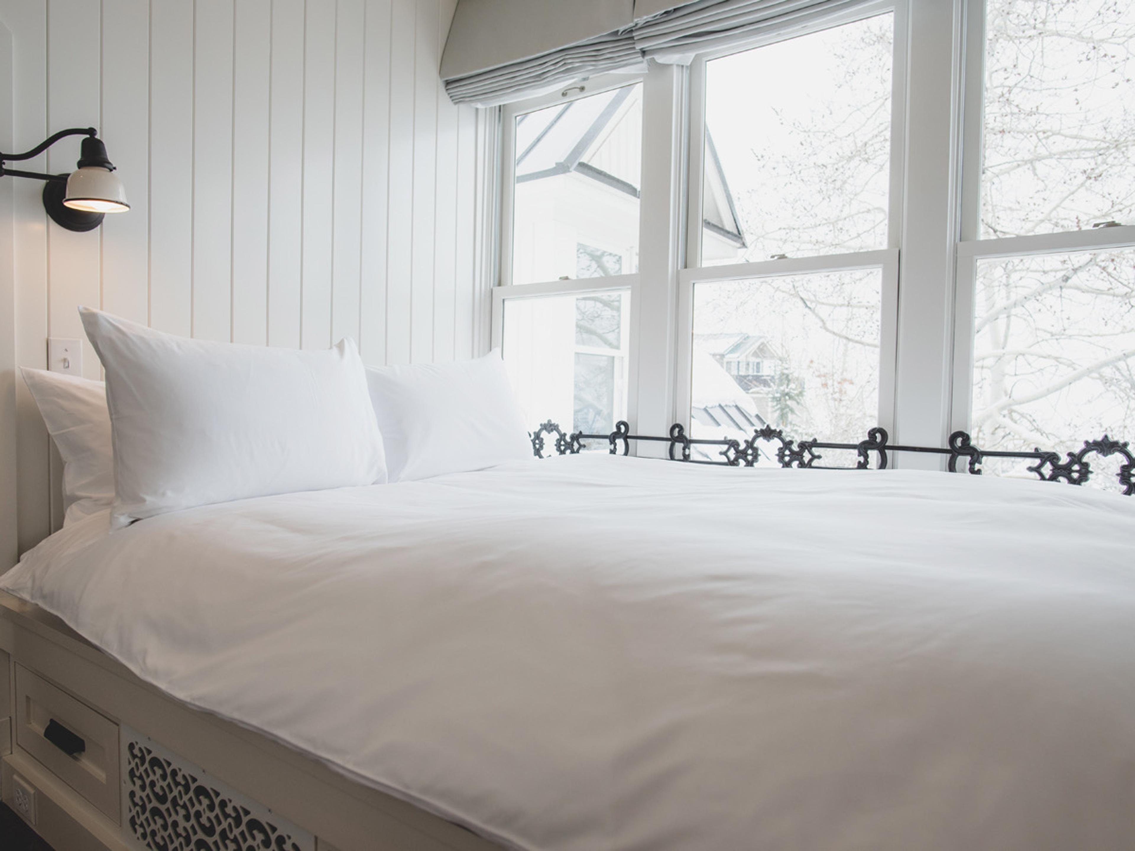 A made bed with white linens in the foreground, facing a window that looks out onto neighbouring rooftops and bare trees.