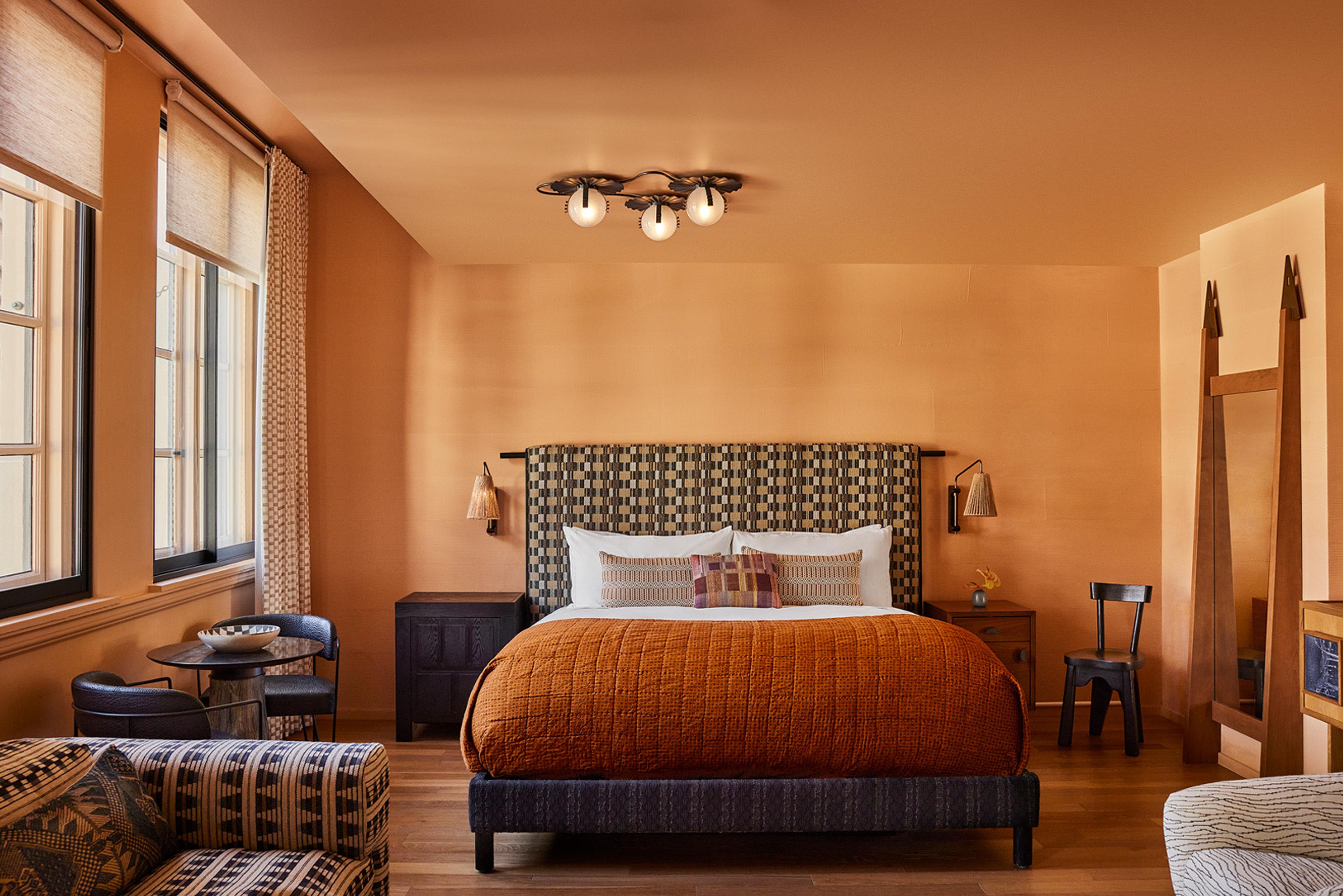 A bedroom with a dark upholstered bed, an orange headboard panel, a rust-colored quilt, and two tall windows framed by a wood ladder and mirror.