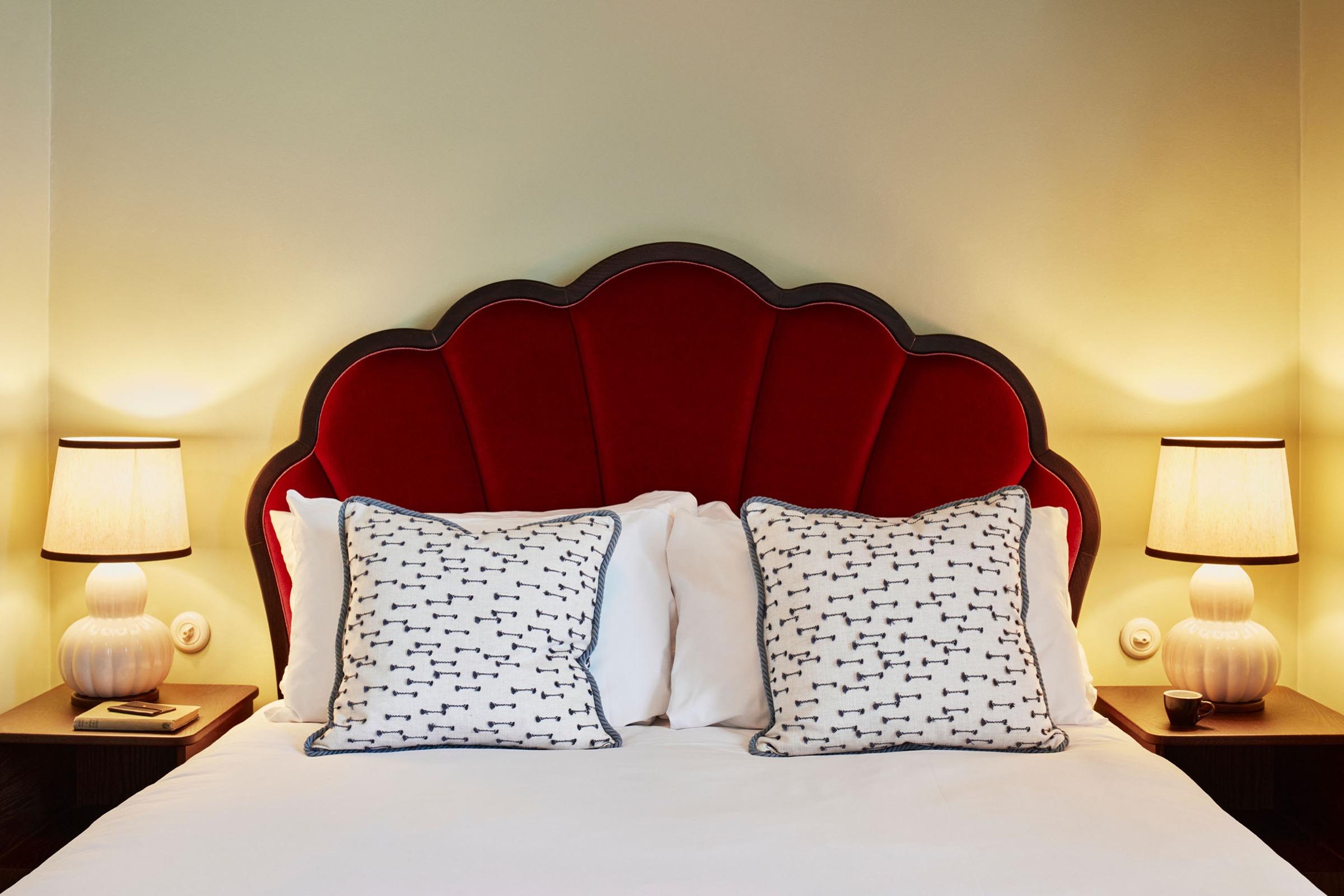 A bed with a scalloped red velvet headboard, patterned cushions and brass reading lamps either side.