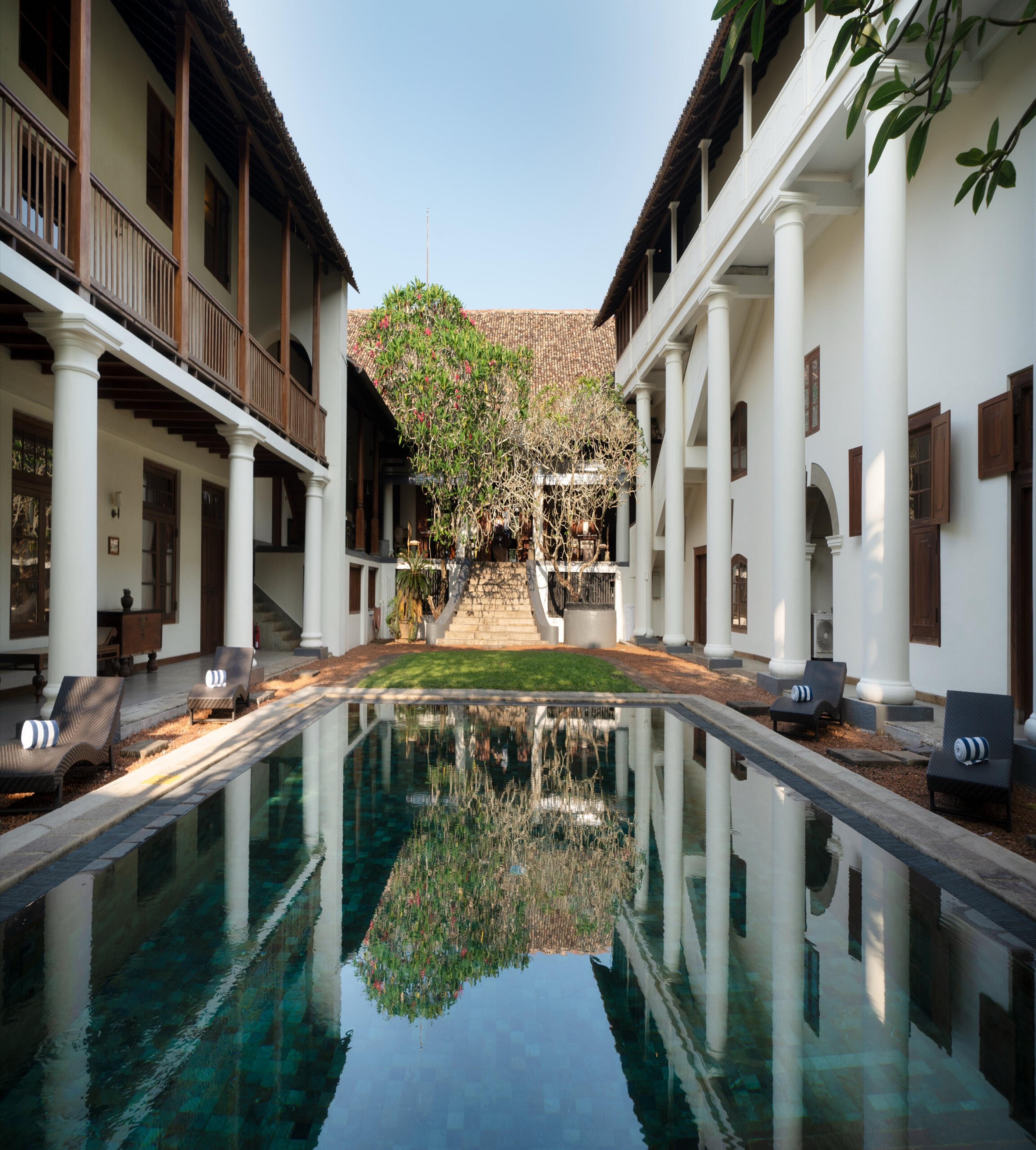 A long reflecting pool down the centre of a colonnaded courtyard, with white pillared buildings either side and a tree and staircase at the far end.