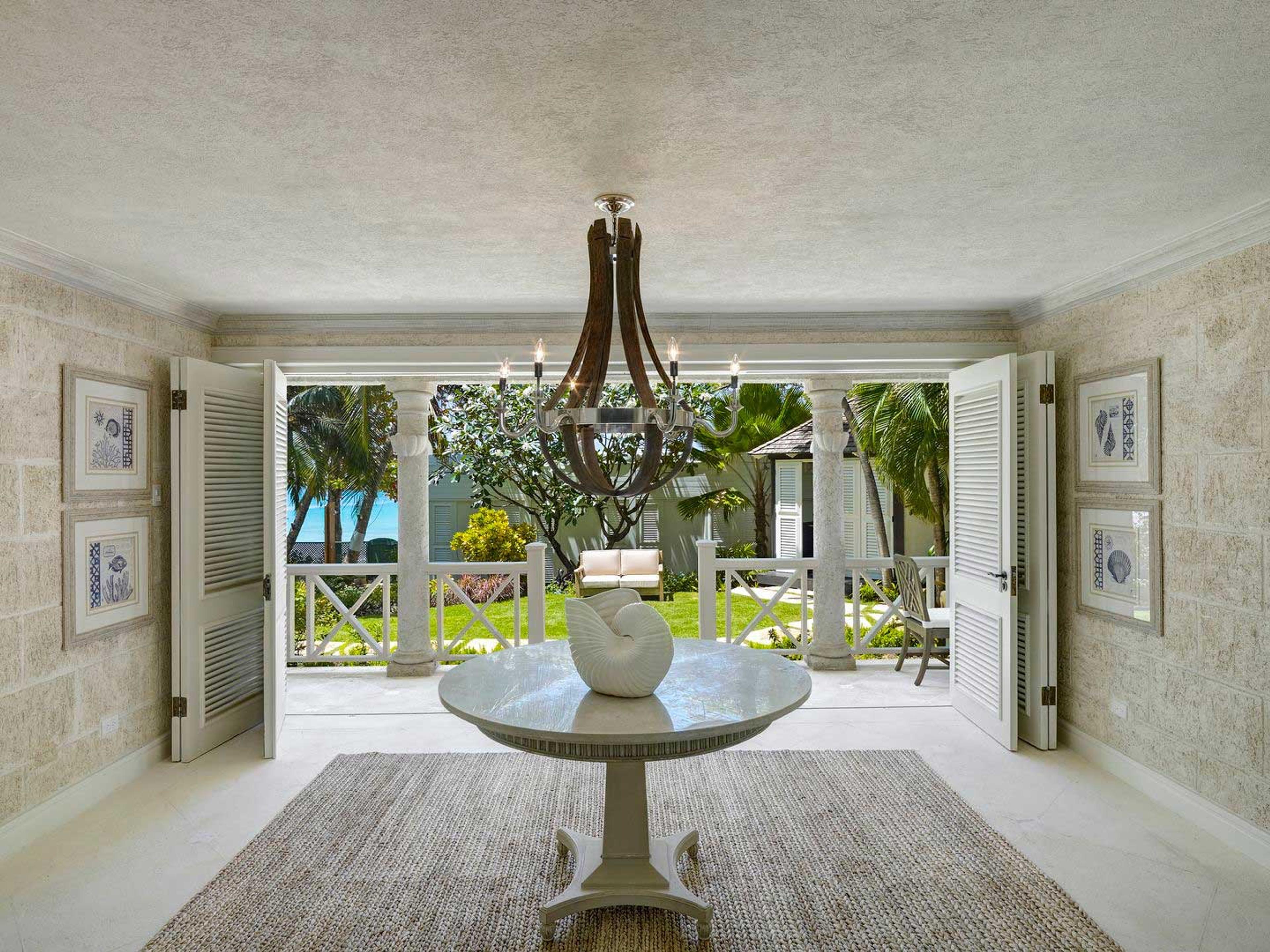 An entrance hall with an iron chandelier over a round table holding a white shell, its shuttered doors open onto a columned veranda and garden
