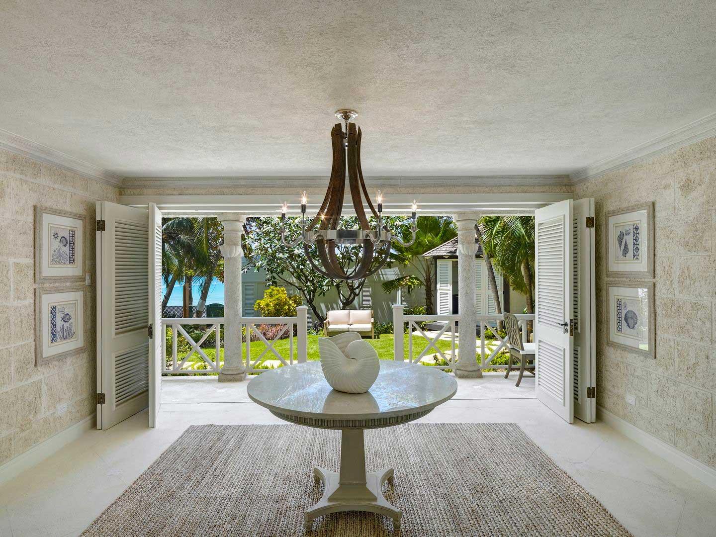 An entrance hall with an iron chandelier over a round table holding a white shell, its shuttered doors open onto a columned veranda and garden