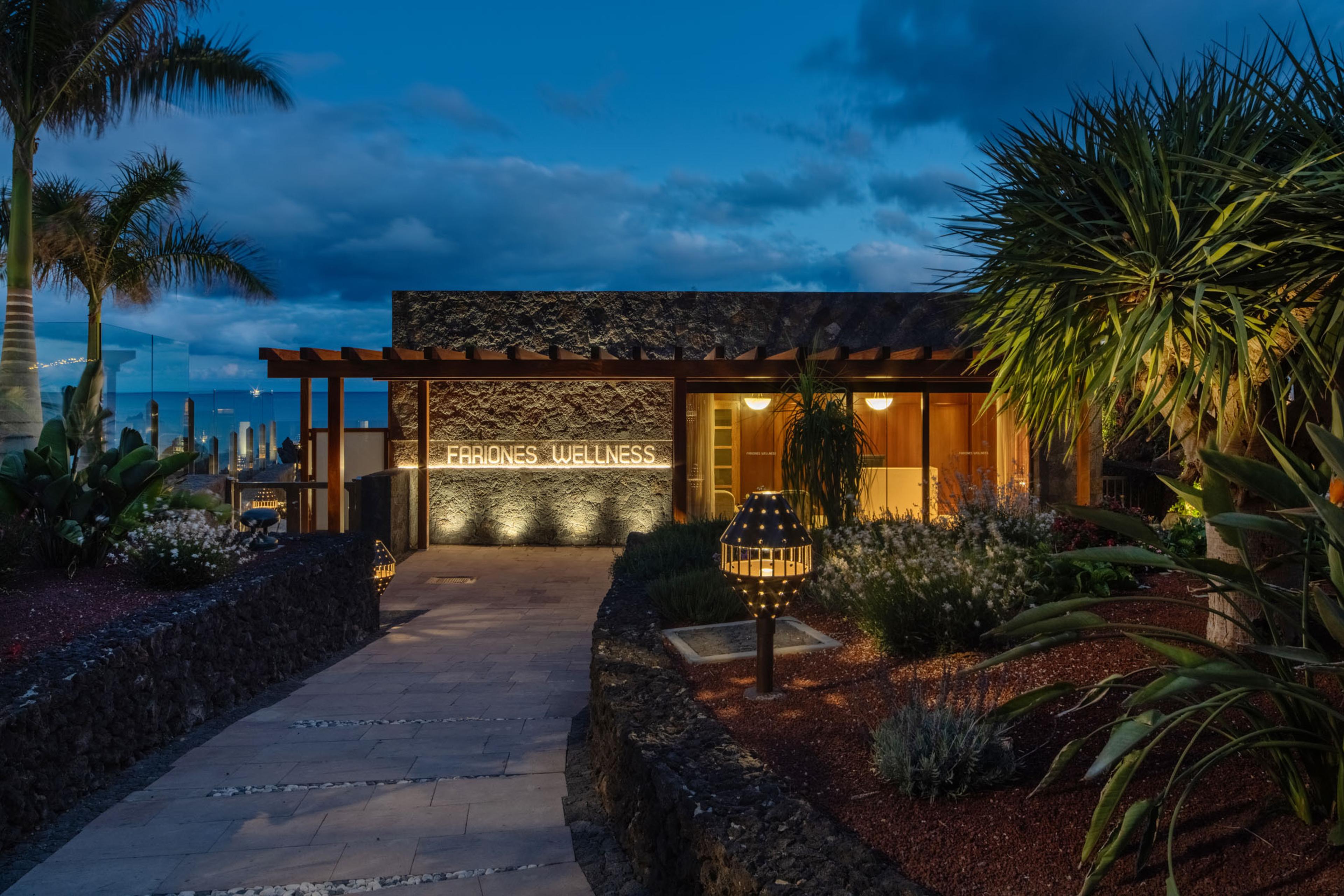 A stone wellness pavilion lit up at night, a sign reading Fariones Wellness glowing beside a pergola, palm trees and a lantern lit path in front.