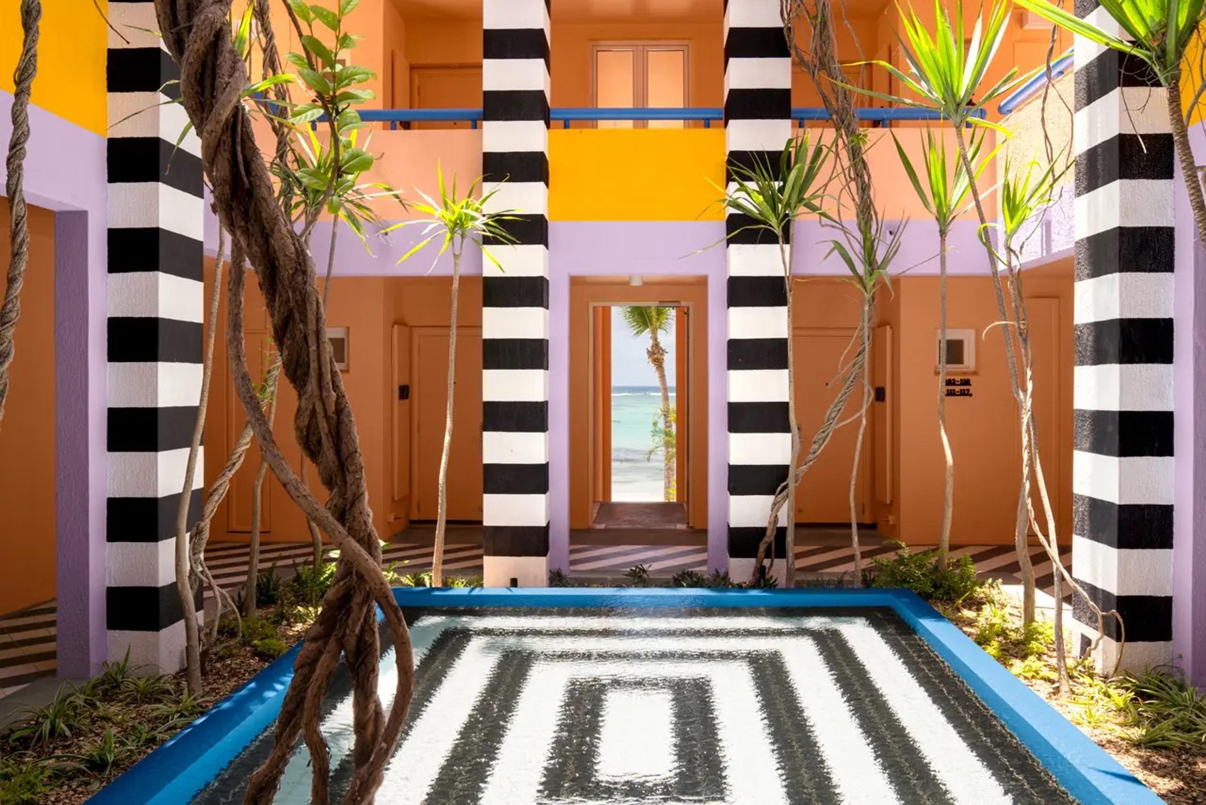 Black and white striped columns surround a tiled plunge pool in a courtyard, framing a doorway that opens onto the beach.