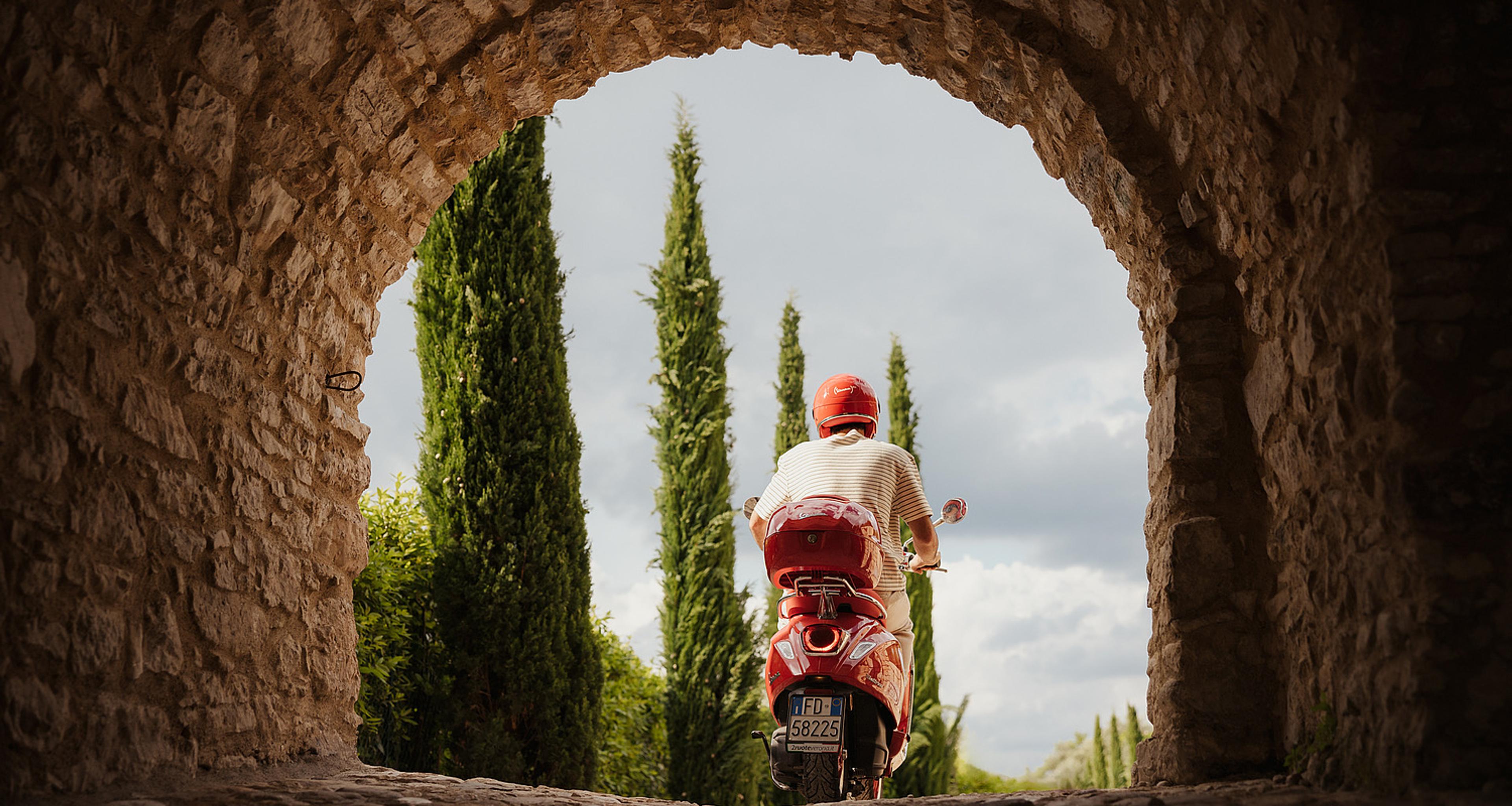A person on a red scooter riding away under a stone archway, cypress trees lining the road ahead.