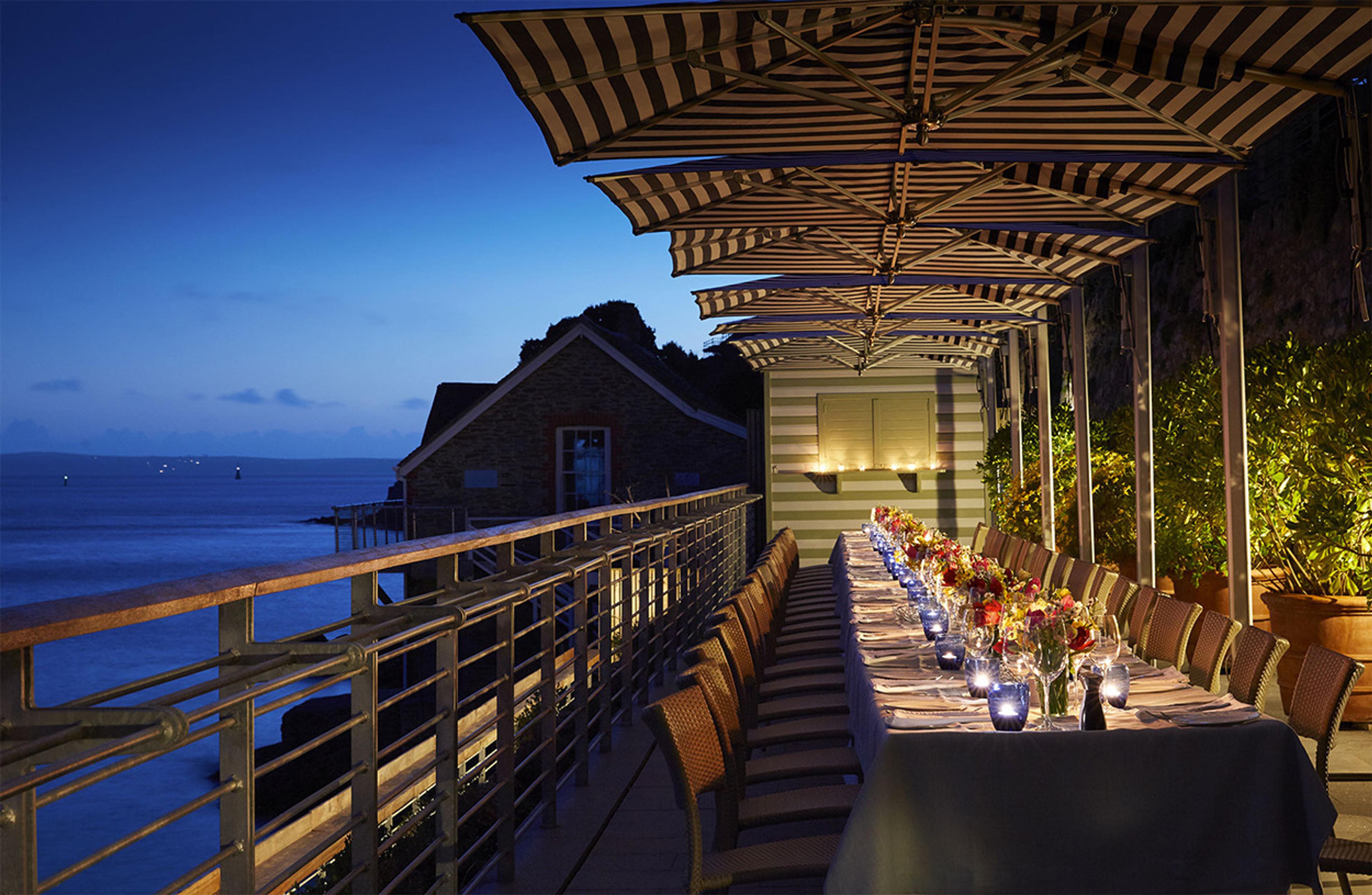 A long outdoor dining table set for many guests beneath striped umbrellas on a terrace above the sea at dusk, with a cottage and dark headland beyond.