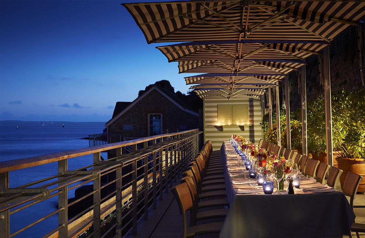 A long outdoor dining table set for many guests beneath striped umbrellas on a terrace above the sea at dusk, with a cottage and dark headland beyond.