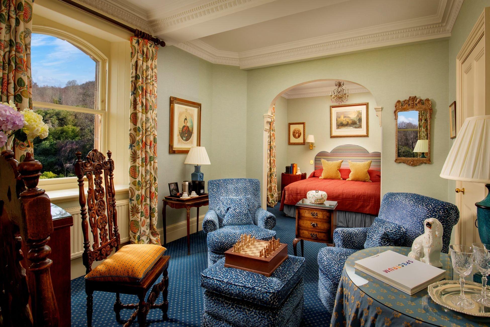 A hotel suite with blue patterned armchairs, a chess set, and an archway opening onto a bed with a red throw and yellow cushions.
