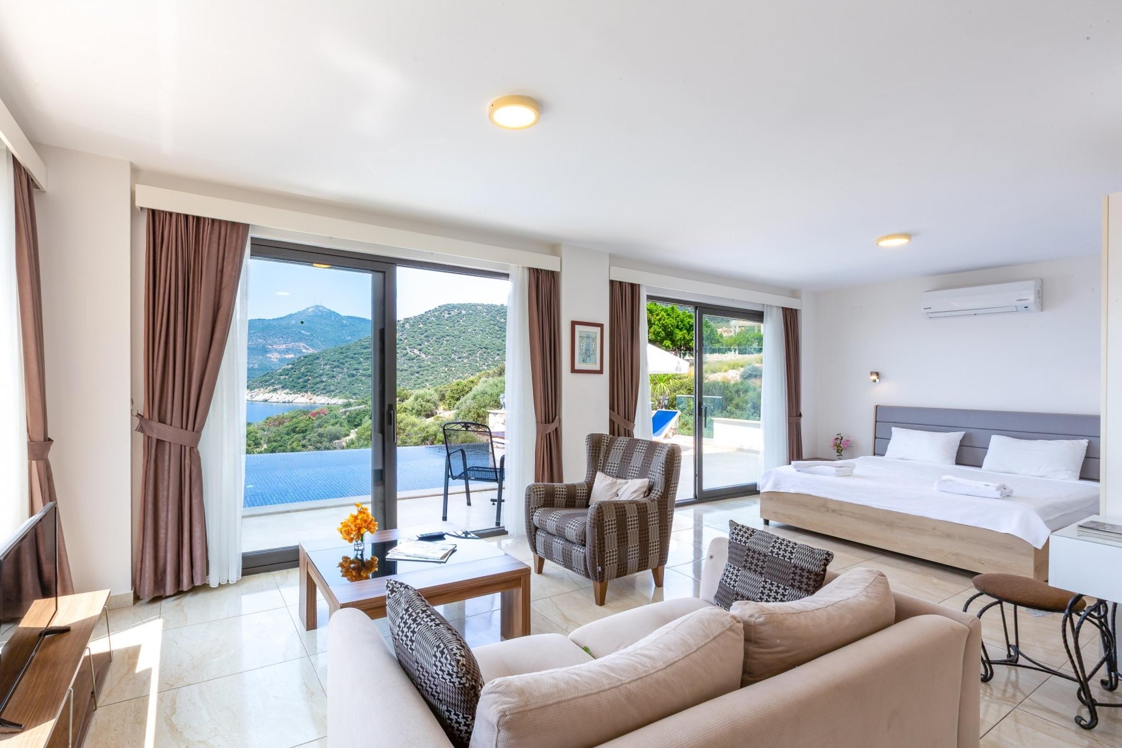 An open-plan hotel suite with a bed, an armchair, and a sofa, with glass doors open onto a balcony that looks over a pool to the sea.