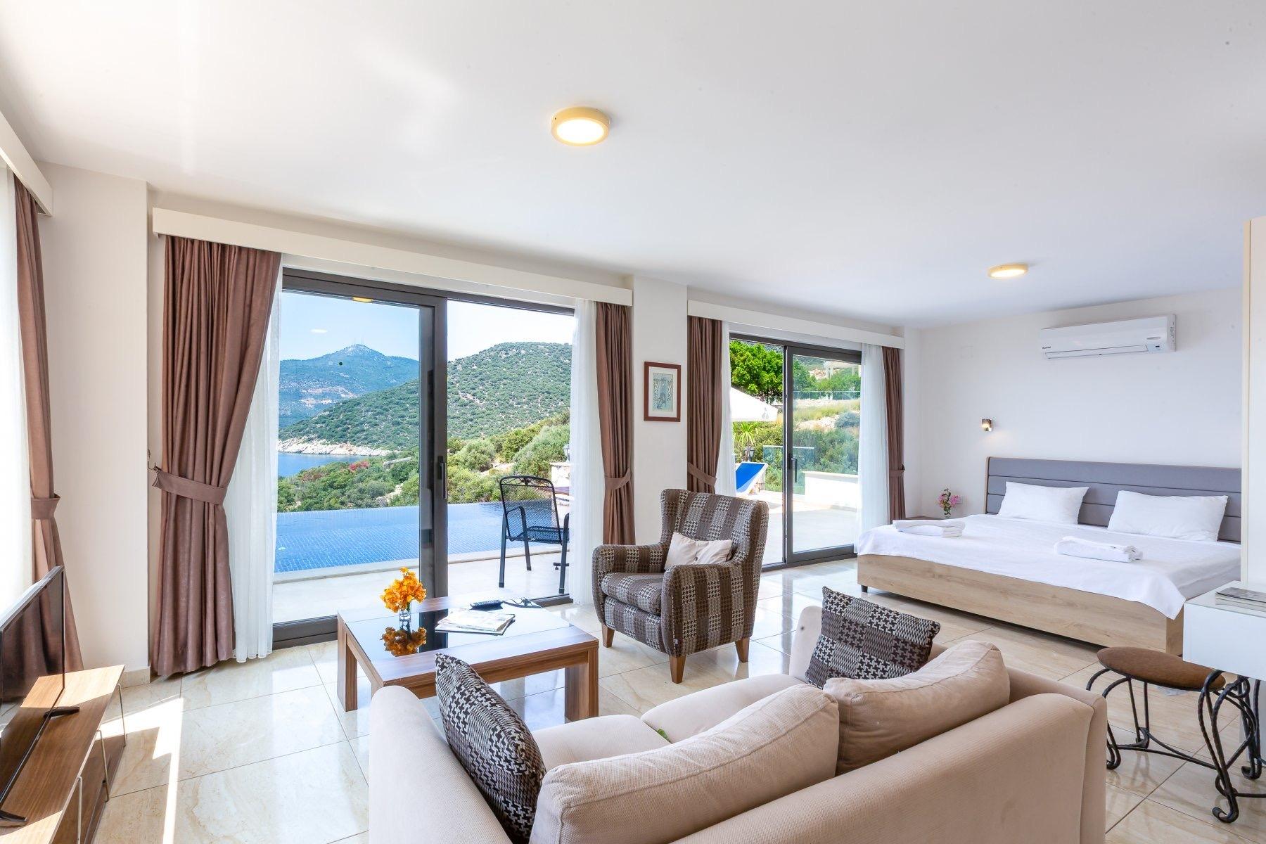 An open-plan hotel suite with a bed, an armchair, and a sofa, with glass doors open onto a balcony that looks over a pool to the sea.