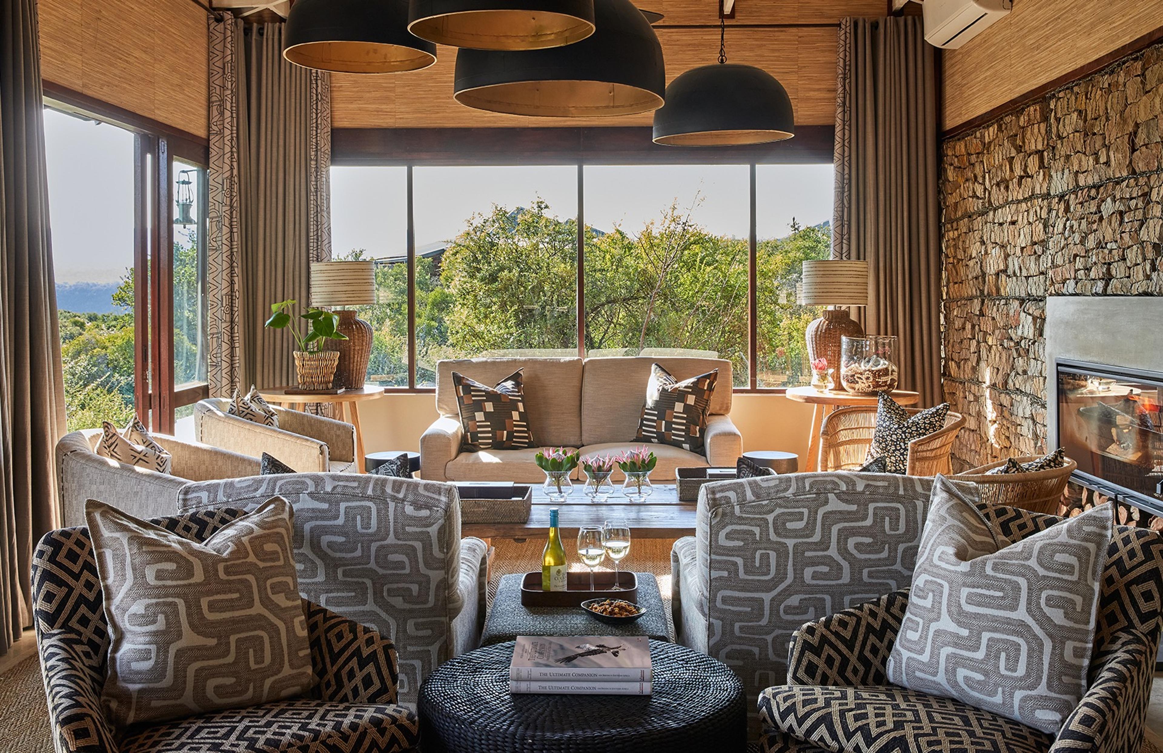 A living room with patterned armchairs, a stone wall fireplace, and tall windows showing trees and hills beyond.