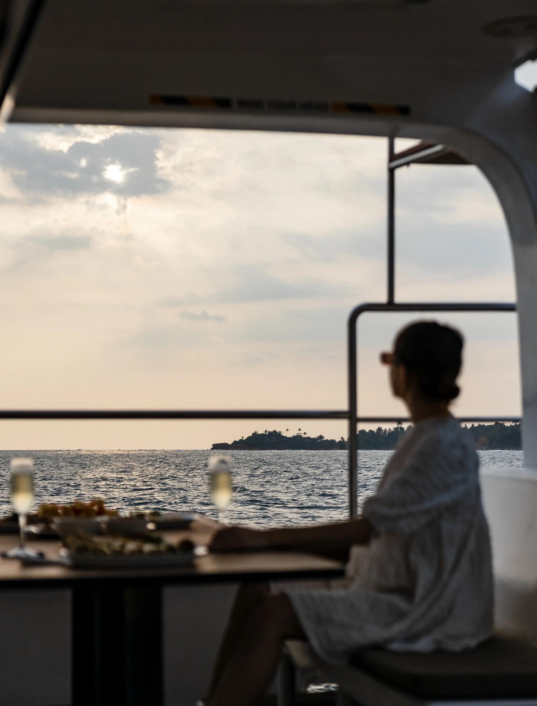 A woman seen from behind on a boat at sunset, with two glasses of champagne and a plate of snacks on the table in front of her.