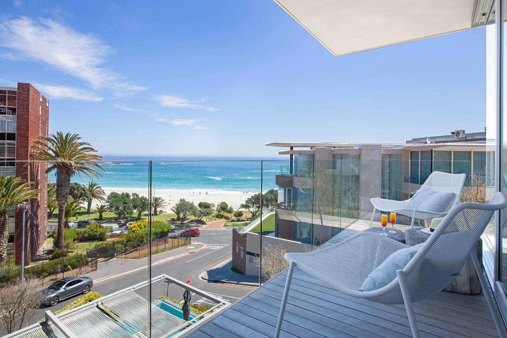 A balcony with pale lounge chairs and a shade umbrella, overlooking Camps Bay beach, the ocean and the coastal road below.