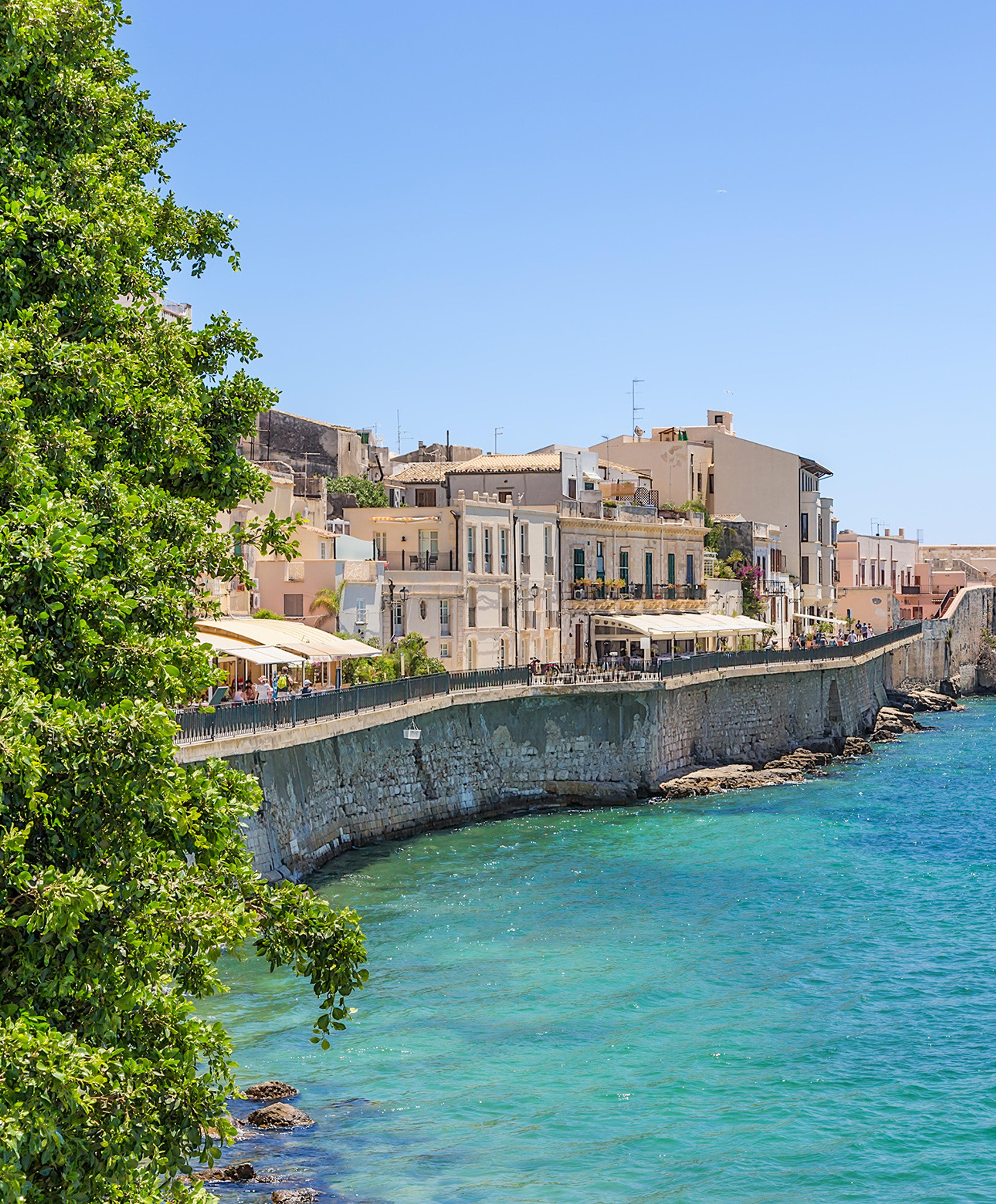 A view along the Ortigia waterfront past a tree in the foreground, with a stone sea wall, pastel buildings and turquoise water under a clear sky.