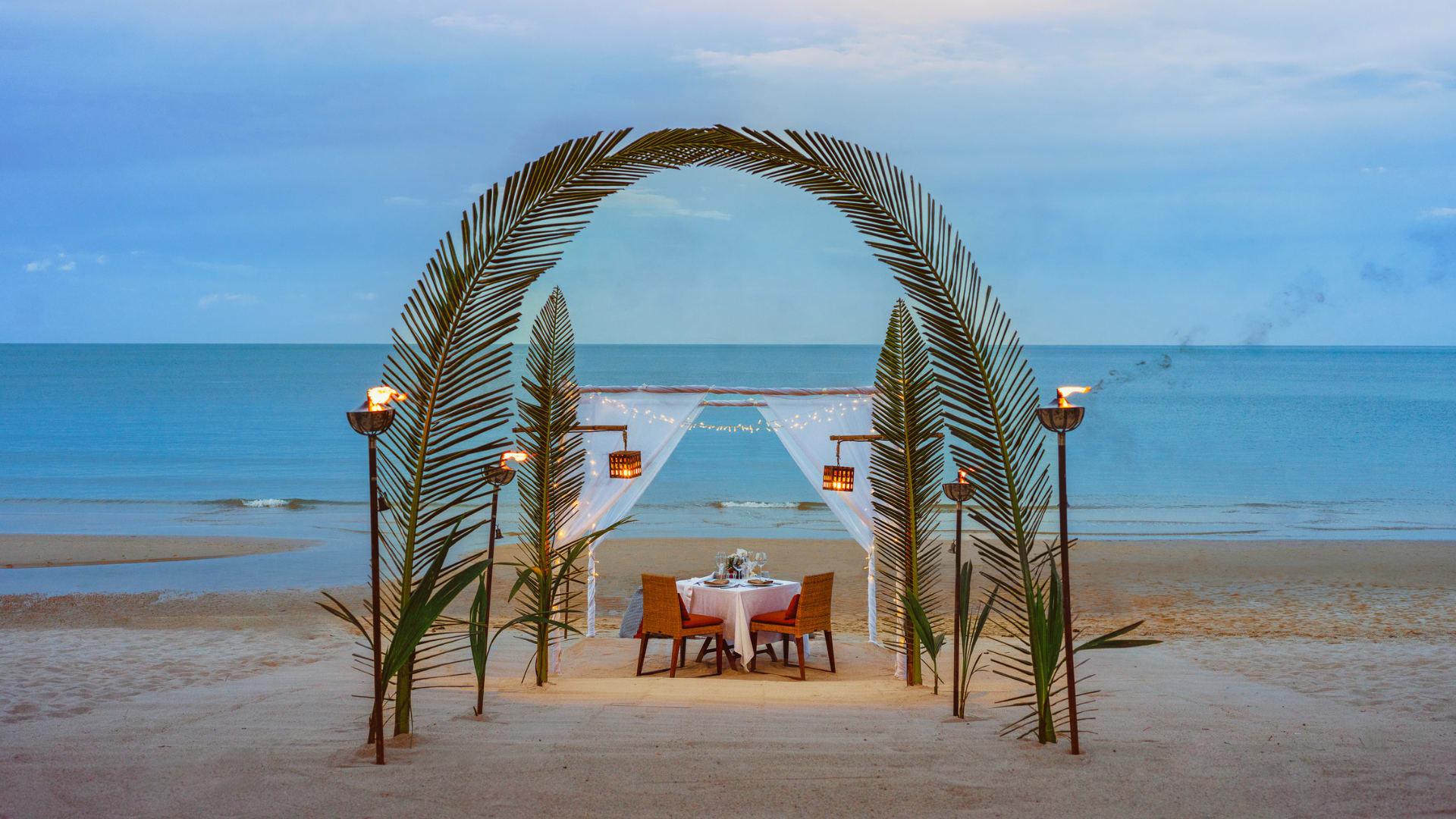 A table set for two under a palm-frond arch on the beach at dusk, lit by torches and string lights, facing the sea.
