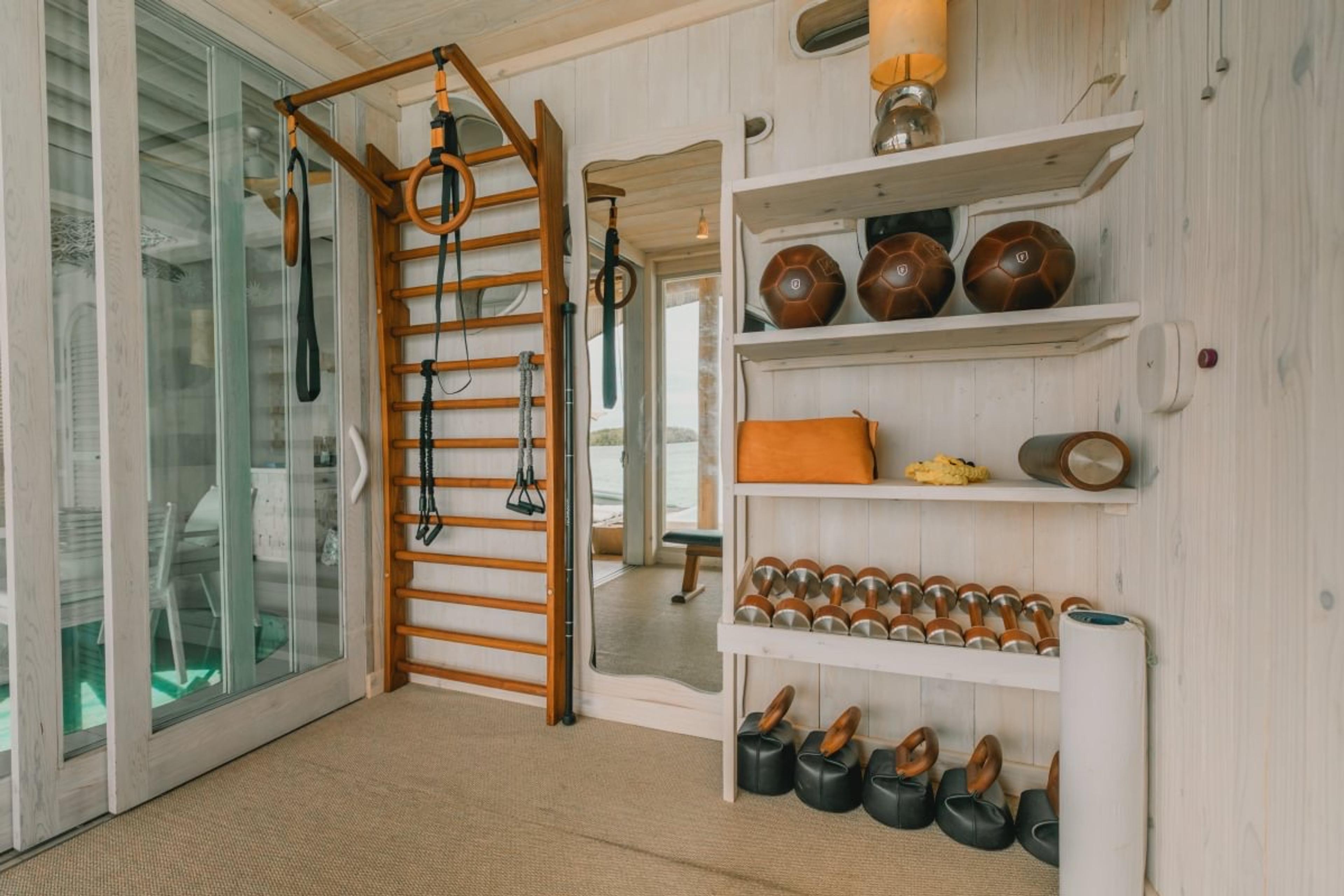 a villa gym with a wooden ladder frame, gymnastic rings, weights and medicine balls on shelves, glass doors open to a deck over the lagoon