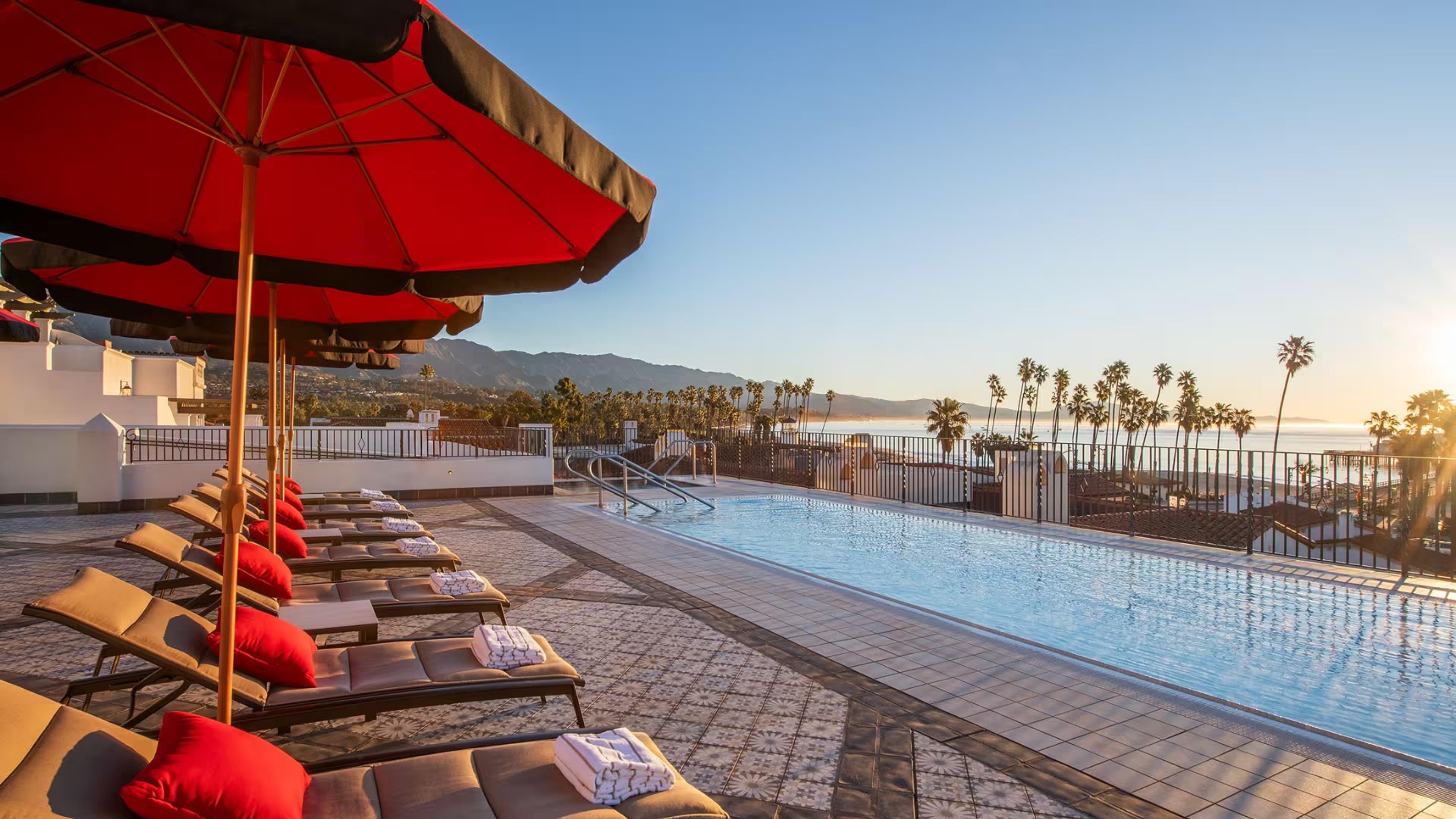 A rooftop pool lined with red cushioned loungers and red umbrellas, with palm trees and the ocean beyond at sunset.