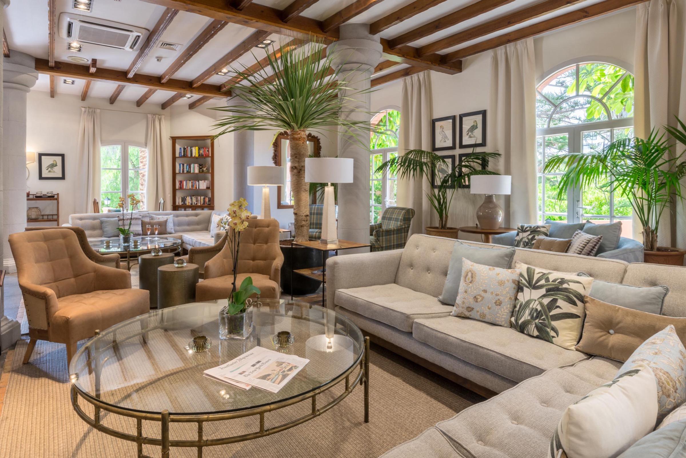 A beamed-ceiling lounge with cream sofas, armchairs, potted palms and tall arched windows looking onto greenery.