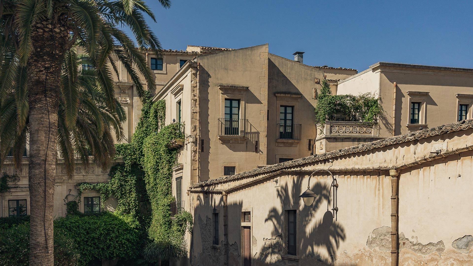 The cream stone facade of a multi-storey palazzo with shuttered windows, ivy climbing one wing, seen past a palm tree under a clear sky.