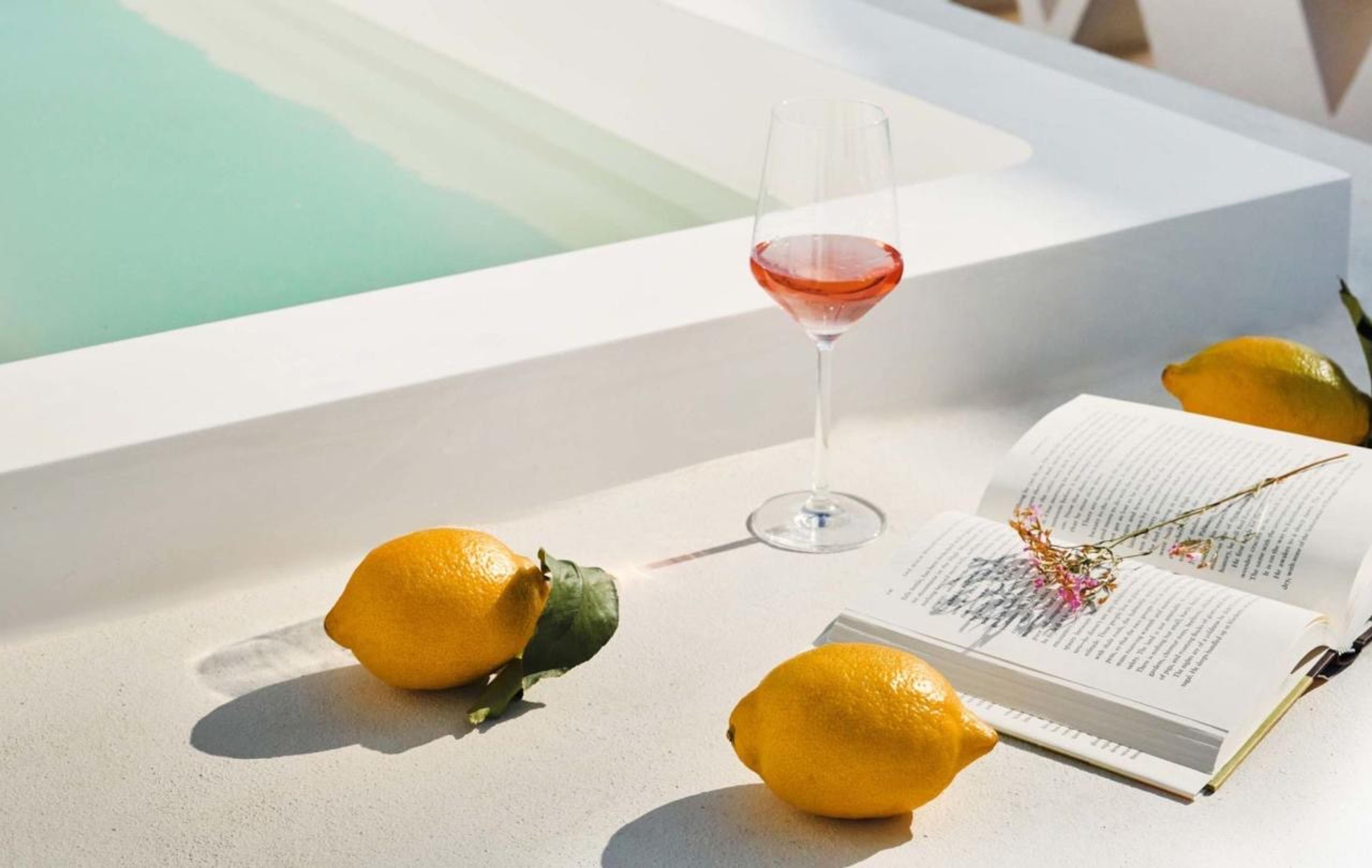 A glass of rosé wine, three lemons and an open book on a white poolside ledge, with turquoise pool water alongside.