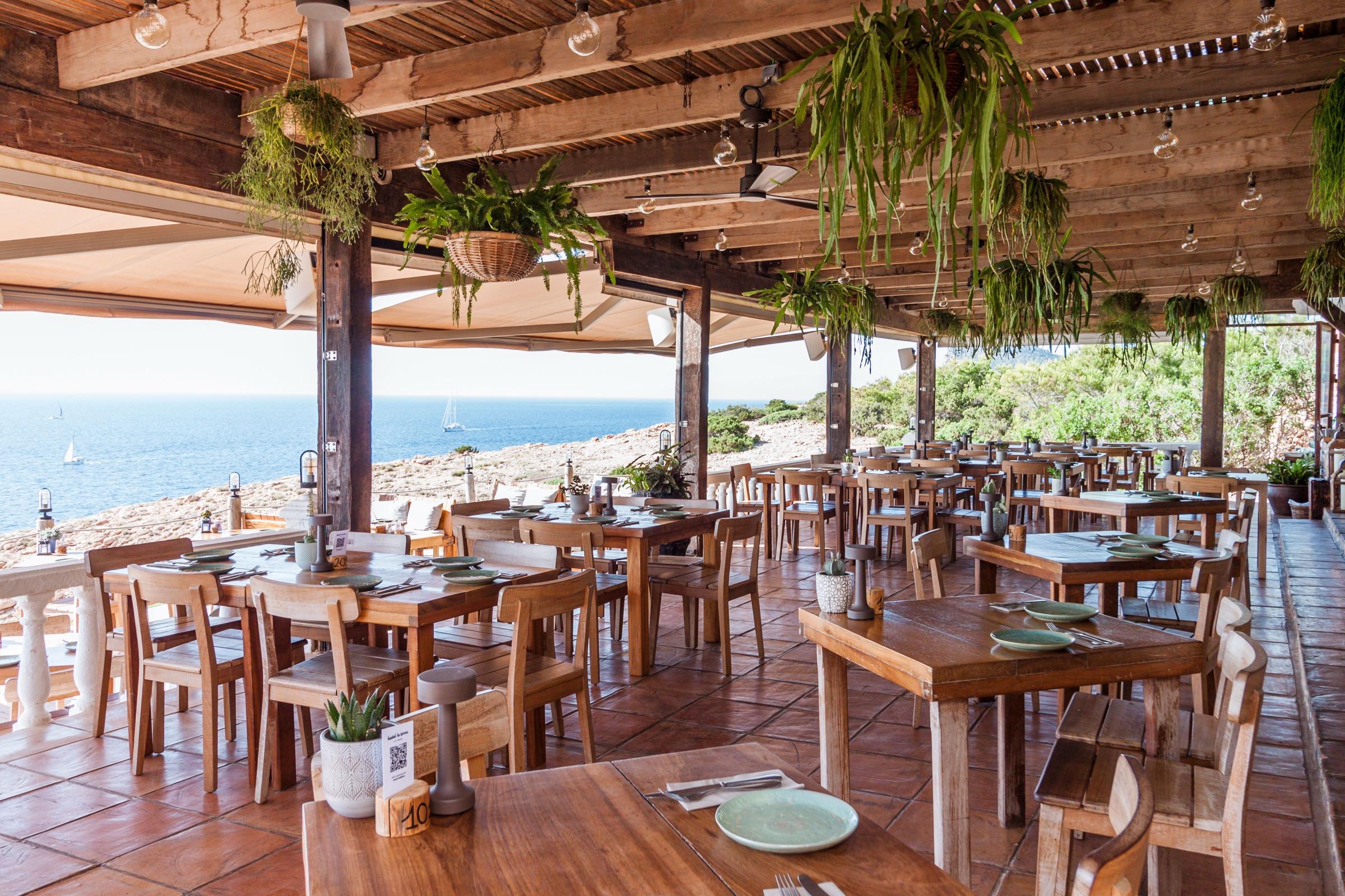 The terrace under a timber pergola hung with ferns, its tables laid, with the sea beyond