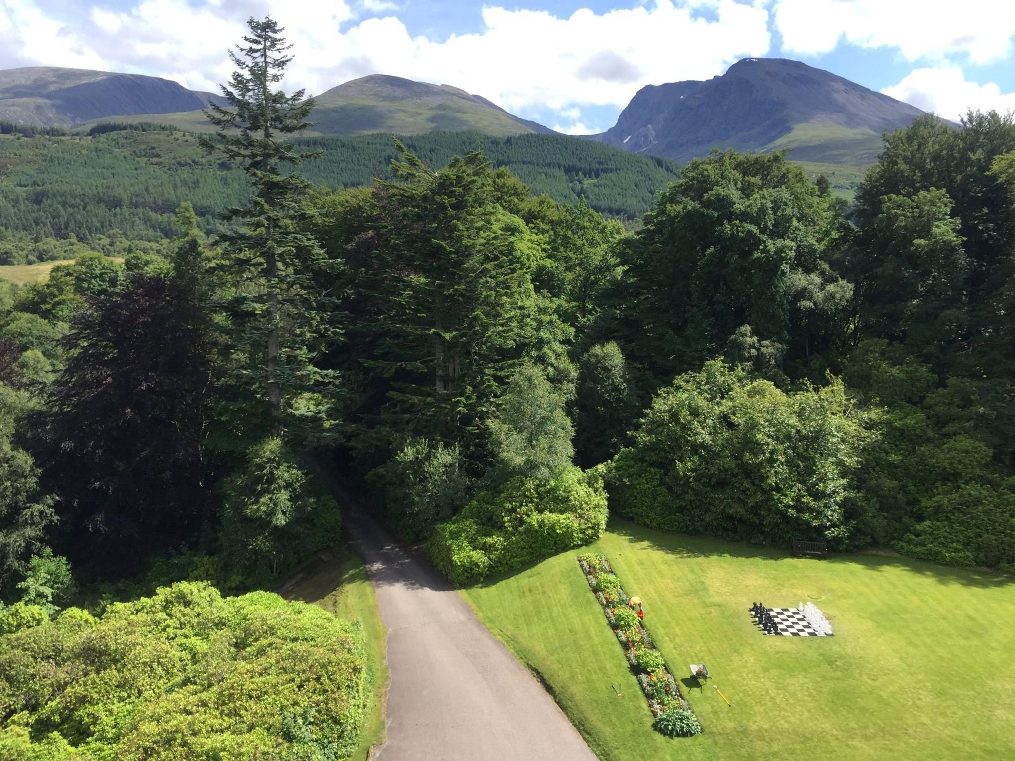 An aerial view of a gravel drive curving through trees toward a lawn with a giant chessboard, mountains behind.