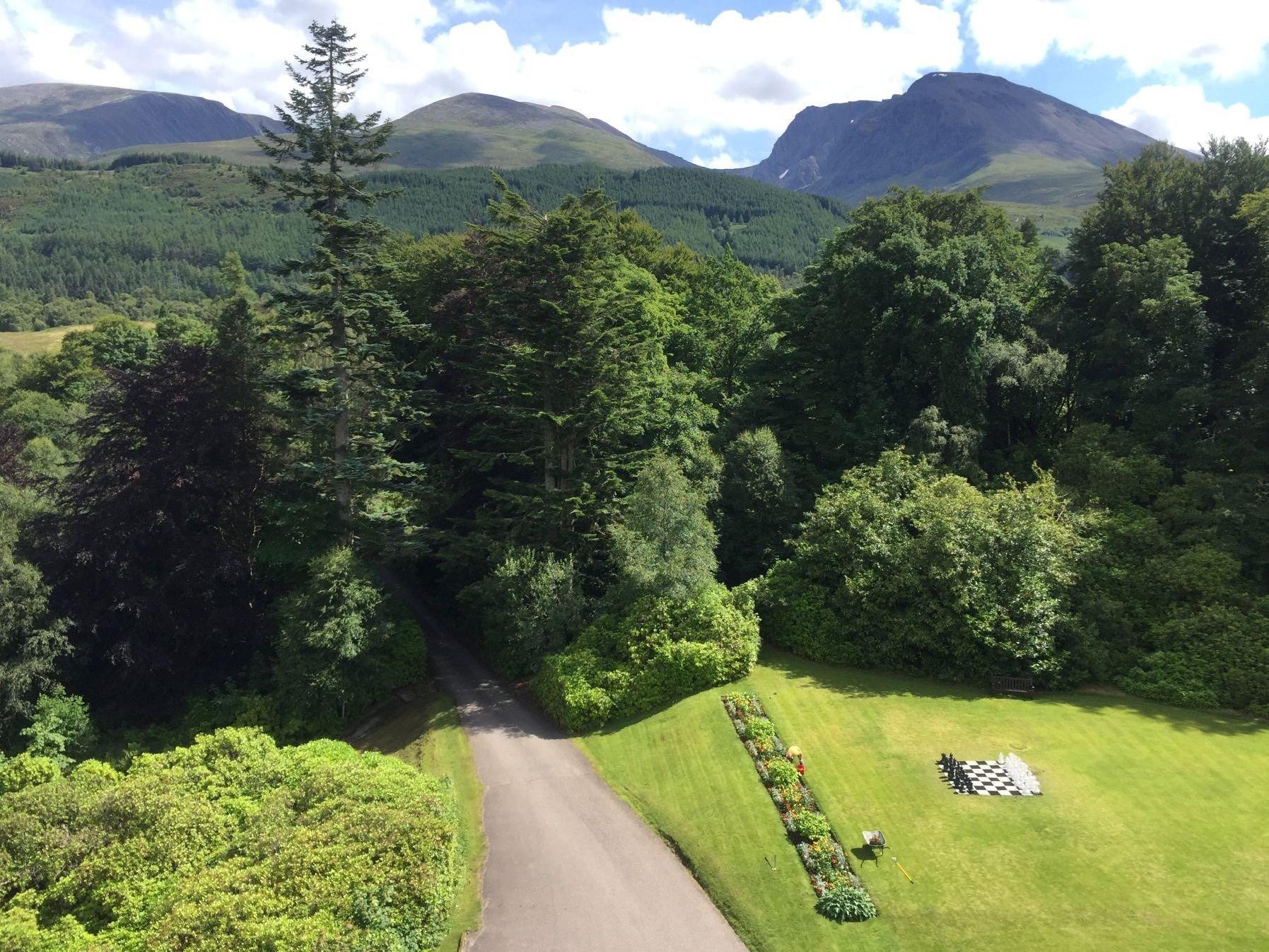 An aerial view of a gravel drive curving through trees toward a lawn with a giant chessboard, mountains behind.