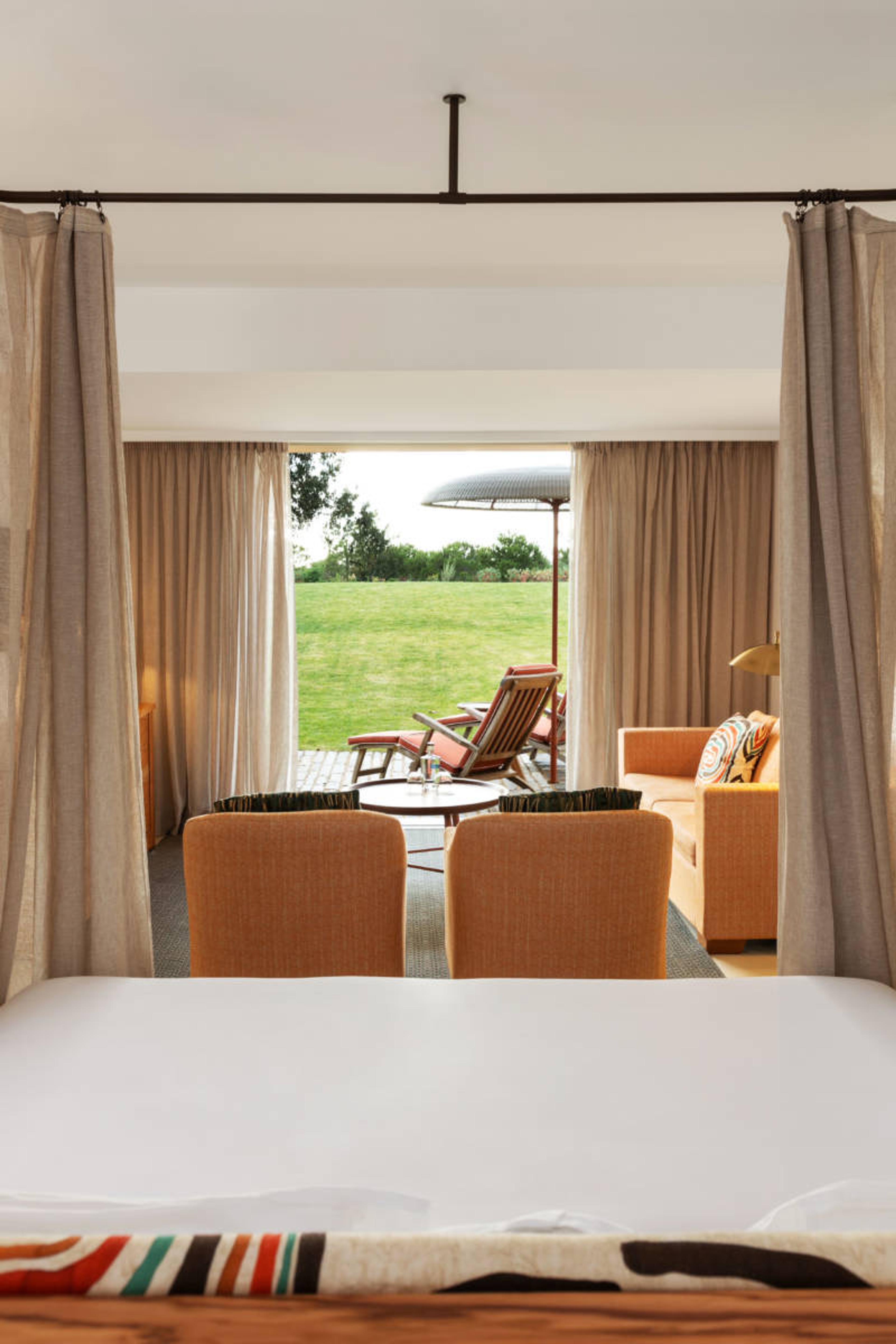 A bed with curtains parted onto a terrace with armchairs, a sofa and an umbrella, lawn beyond.
