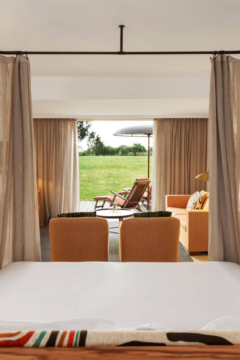 A bed with curtains parted onto a terrace with armchairs, a sofa and an umbrella, lawn beyond.