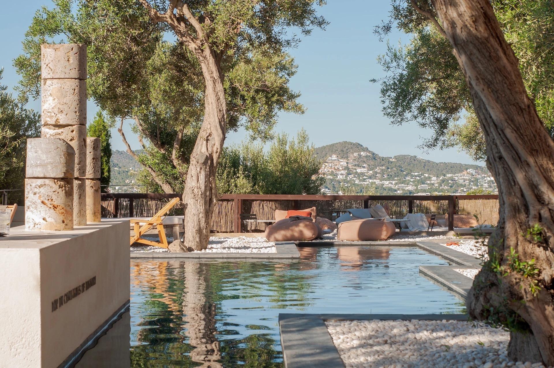 A long pool under olive trees, with stone columns at one end, low cushioned loungers on the pebbles, and a hillside of white houses beyond the wooden fence.