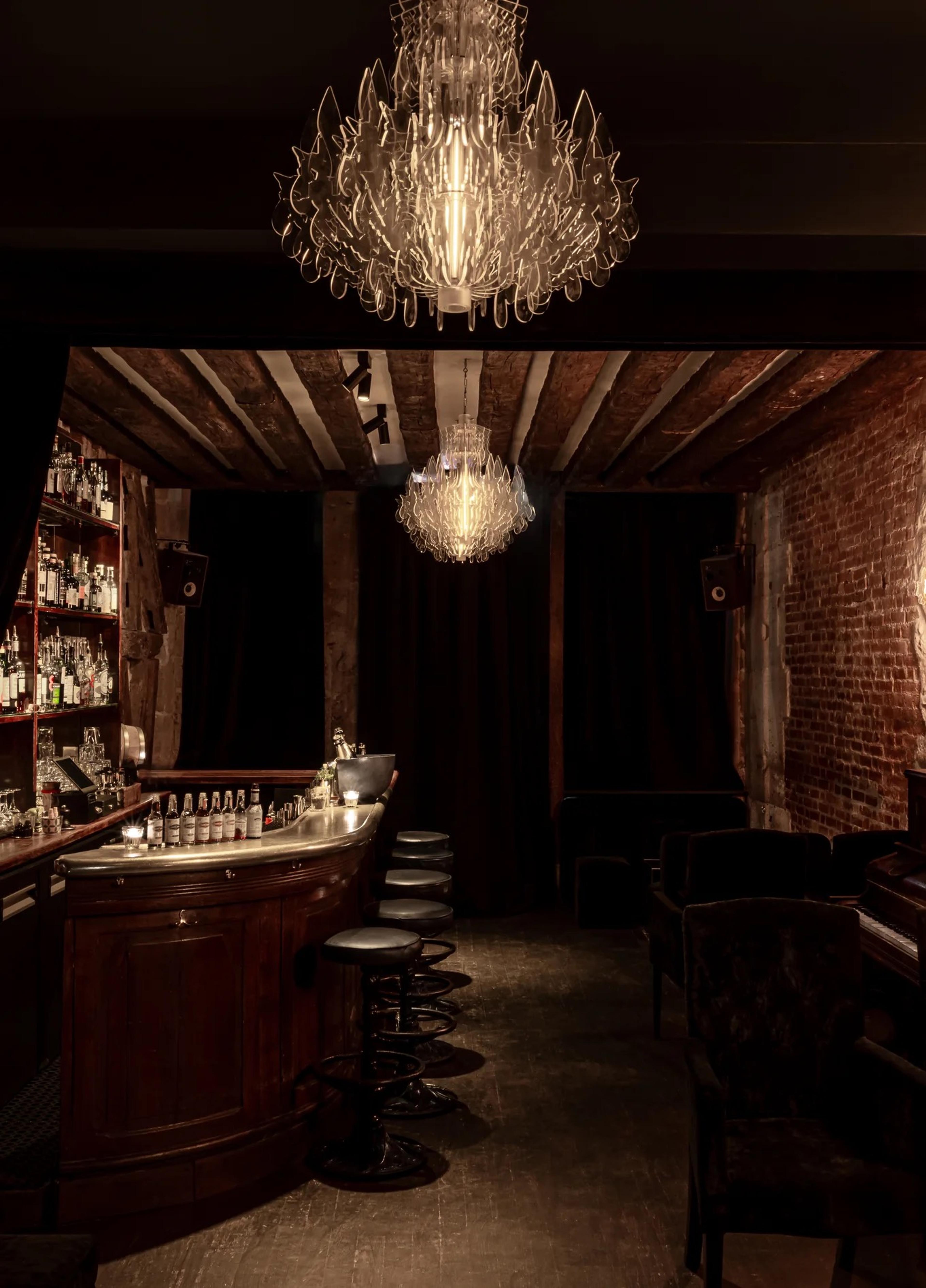 A dimly lit bar with a crystal chandelier, exposed brick walls and timber ceiling beams, bar stools lined along a curved wooden counter.