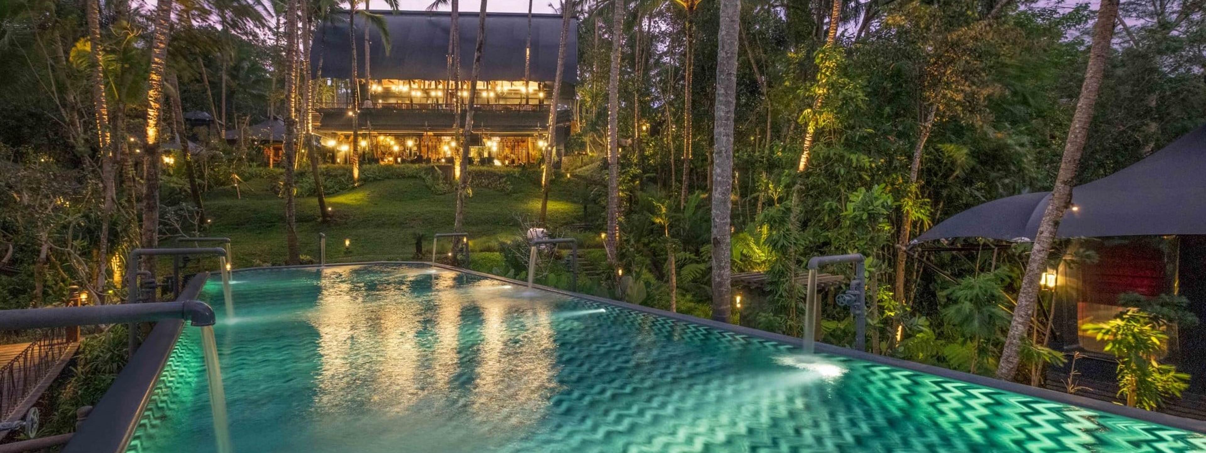 An infinity pool with illuminated water jets in the foreground, and a two-storey open-sided building lit from within beyond it, surrounded by palm trees at dusk.
