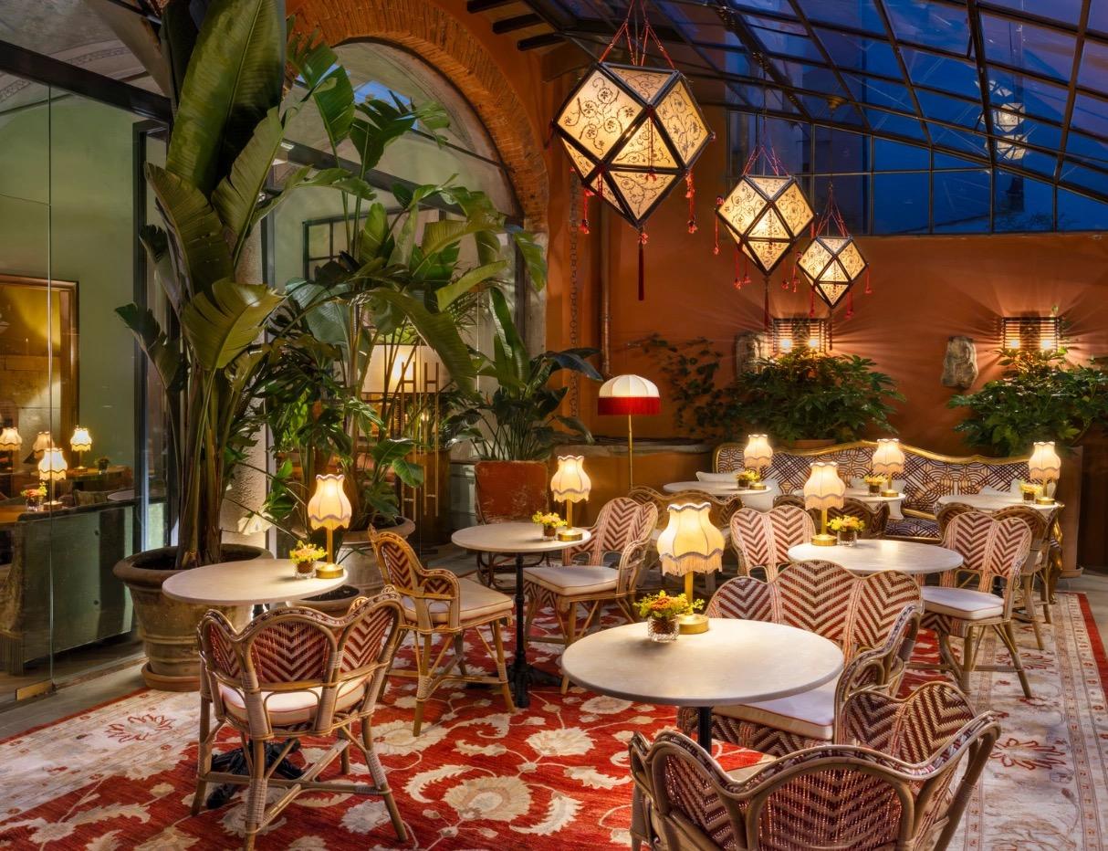 An indoor restaurant under a glass roof, with lanterns hanging over round tables, rattan chairs and tall plants.