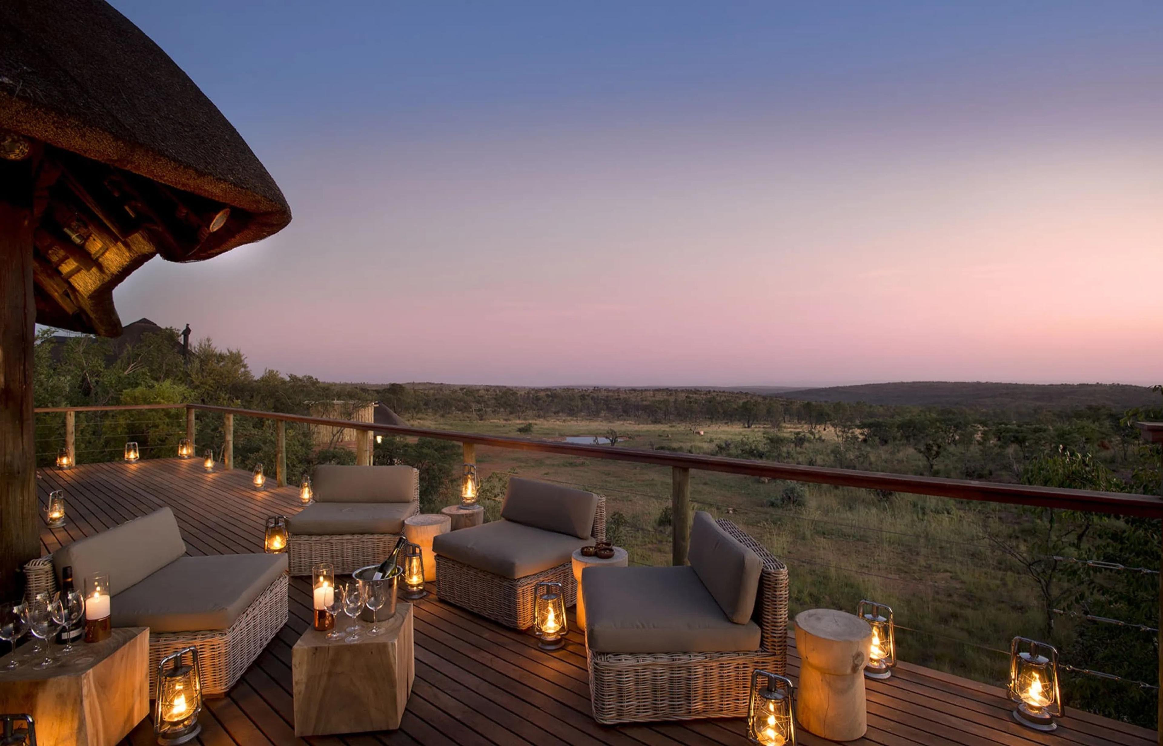 A wooden deck at dusk with wicker armchairs and lantern lights, looking over open bush to a pink sky.
