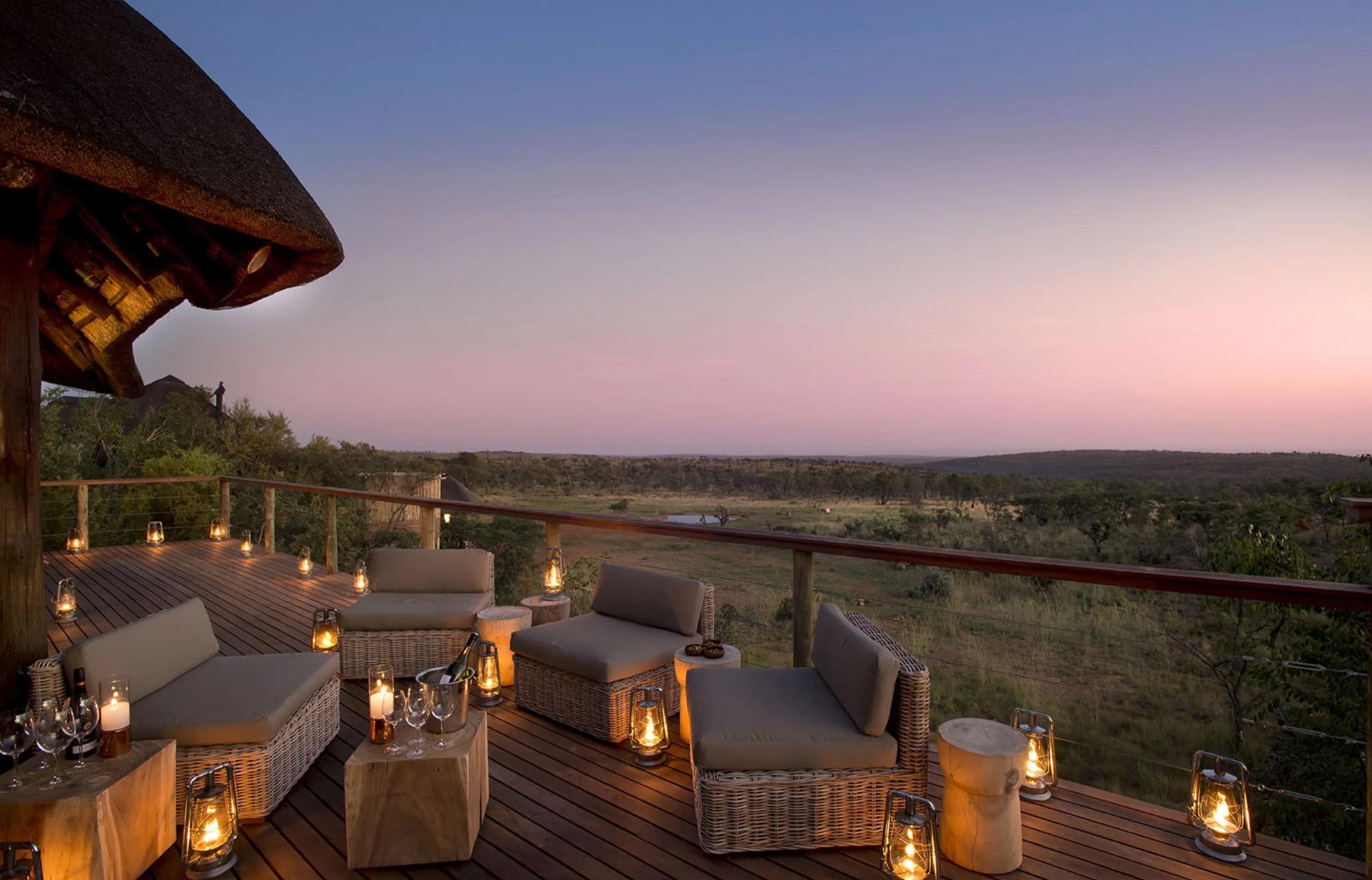 A wooden deck at dusk with wicker armchairs and lantern lights, looking over open bush to a pink sky.