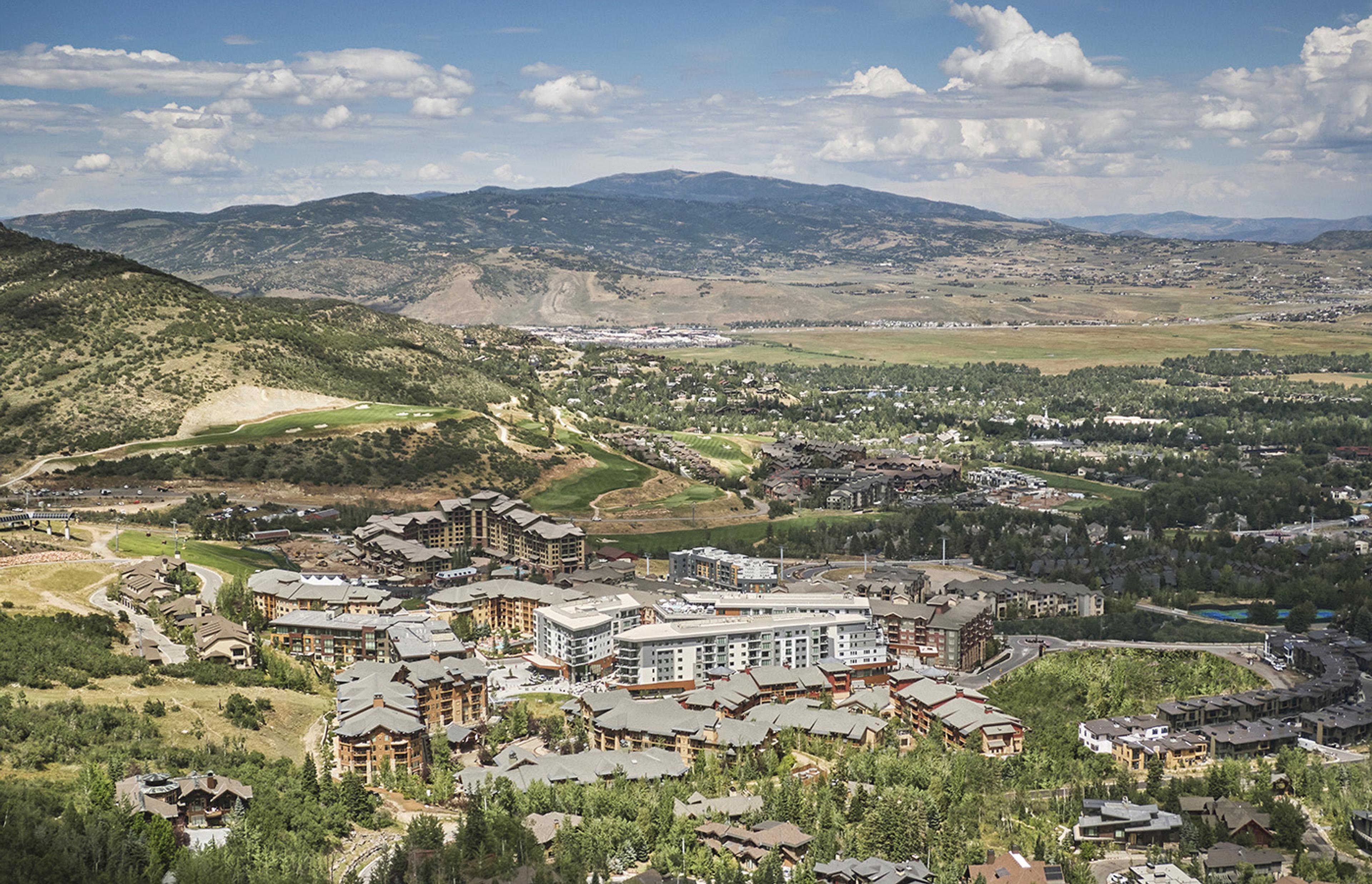 Aerial view across a summer valley toward a hillside, with a cluster of hotel buildings in the middle distance, houses and trees below, and a mountain range beyond.