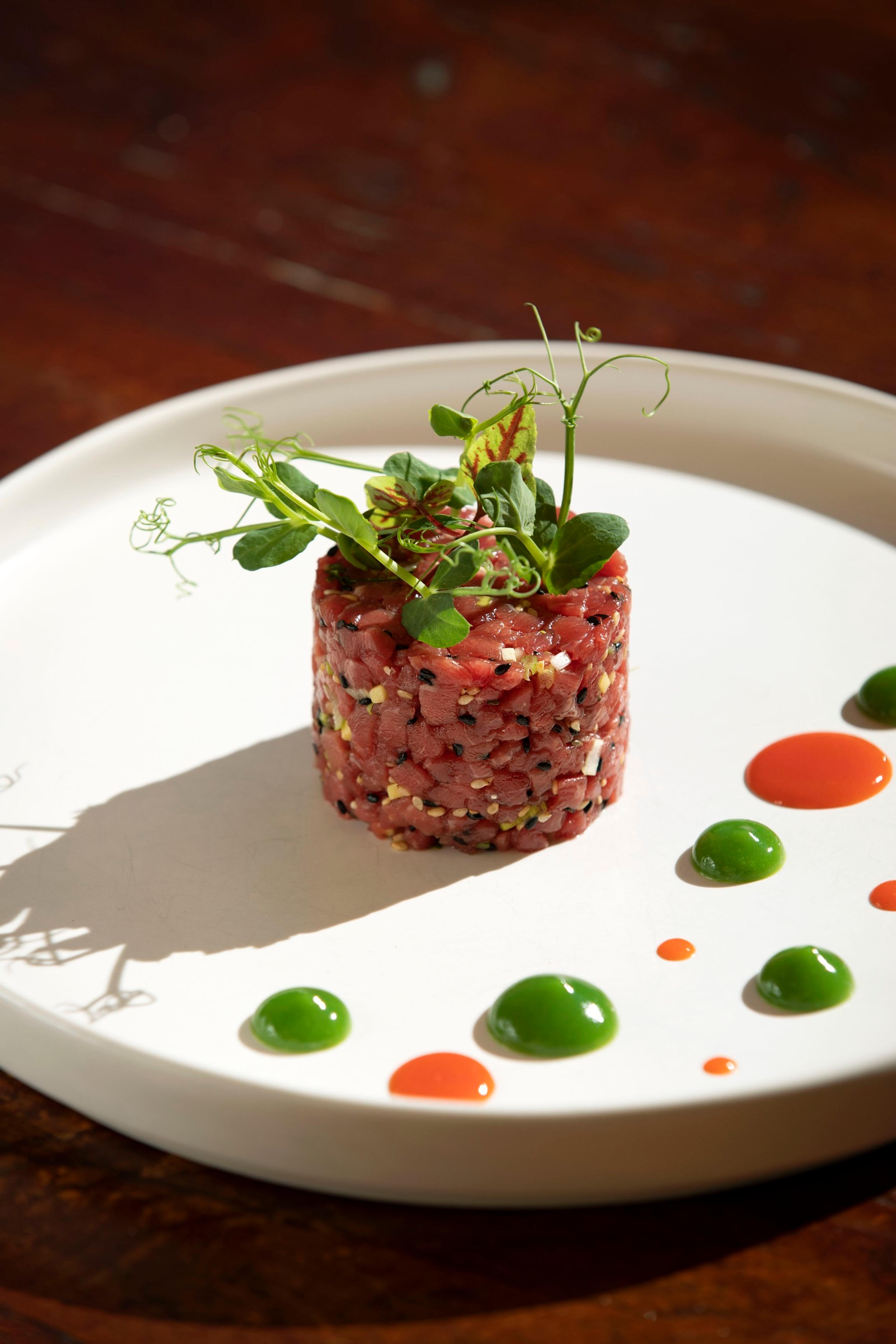 A plate of tuna tartare topped with pea shoots, with small dots of red and green sauce around it.
