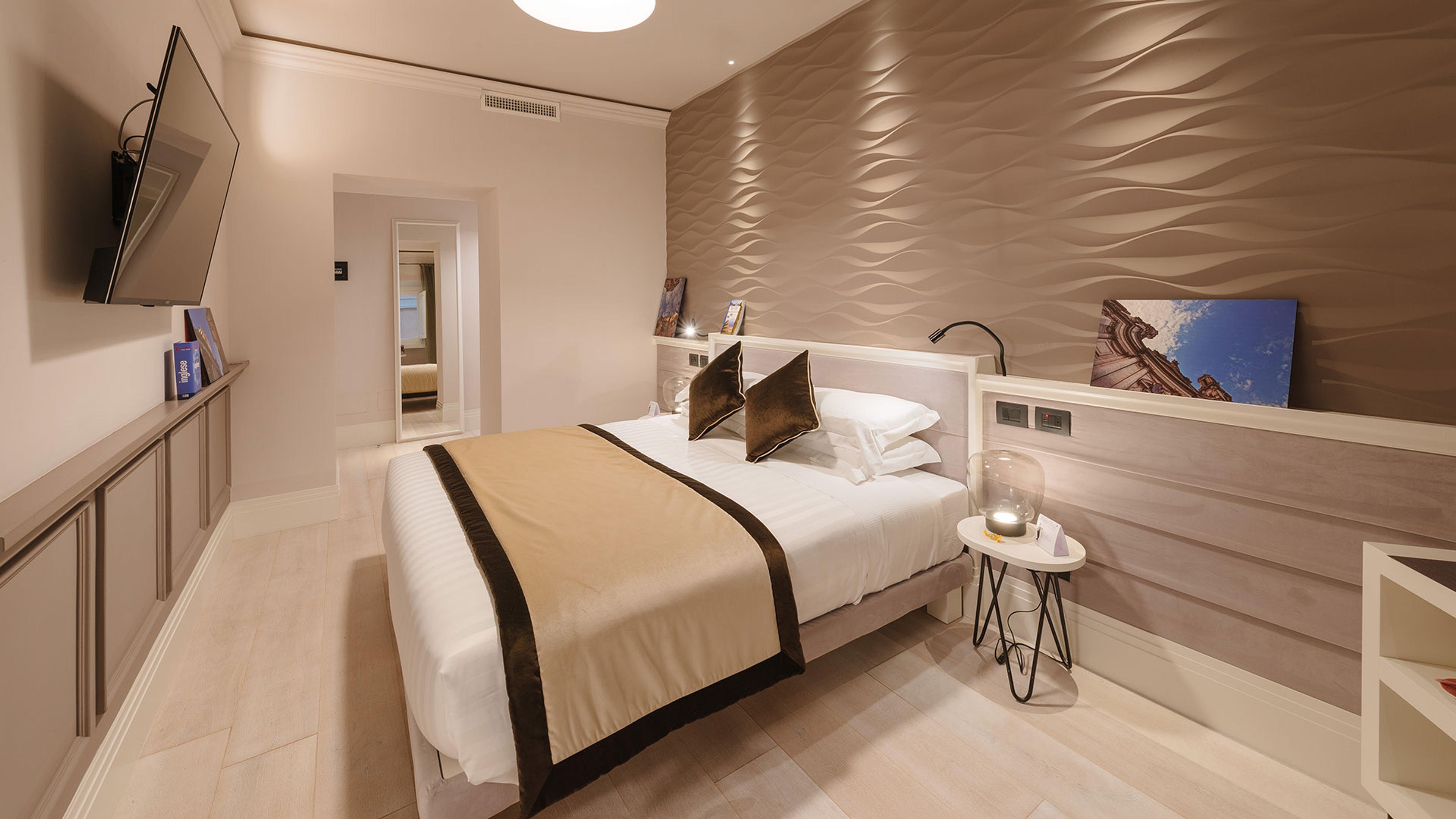 A bedroom with a textured brown wave-pattern wall, a made-up bed with a tan throw, a wall-mounted television over a low sideboard, and a glimpse of a mirrored hallway beyond.