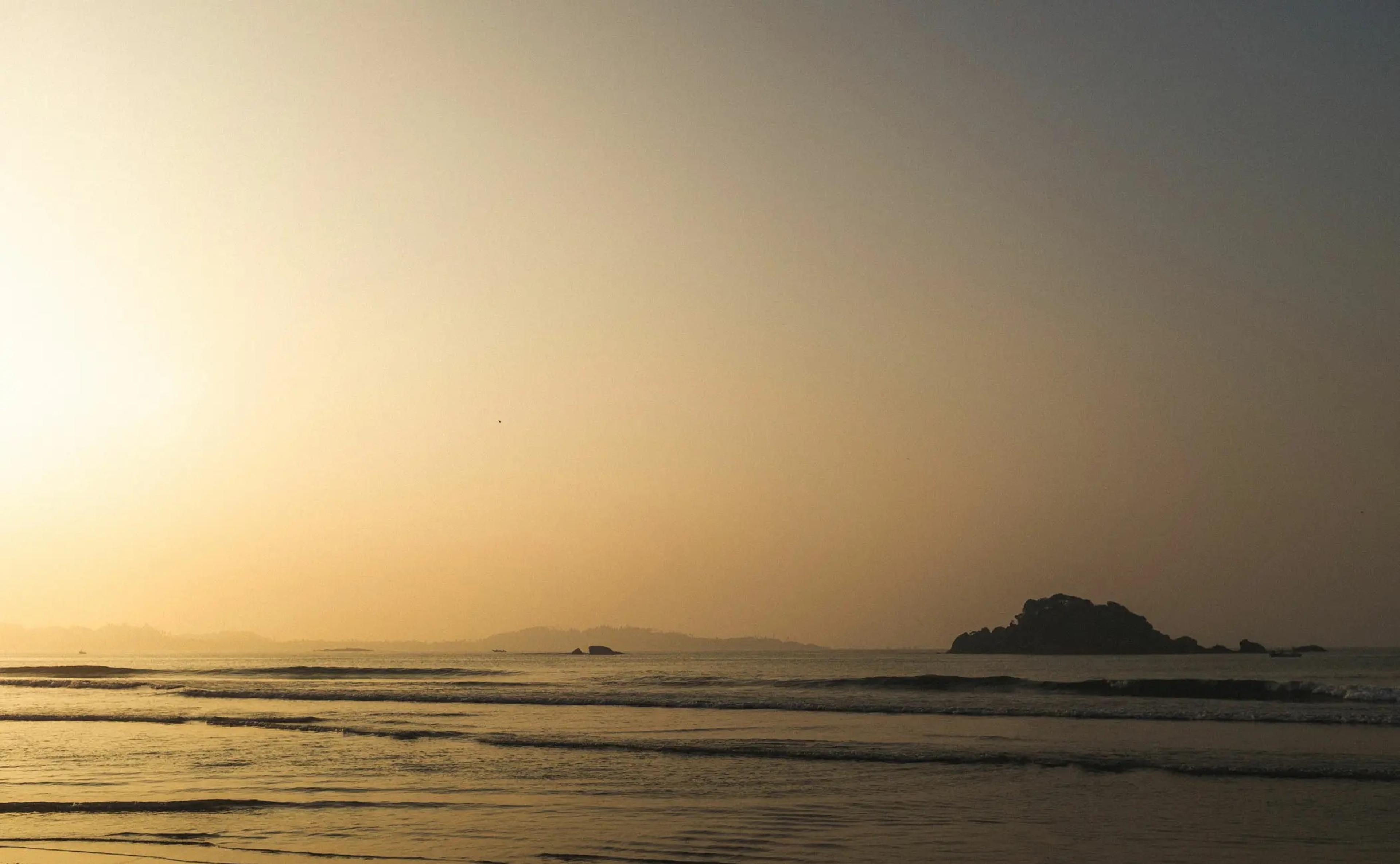 A hazy sunset over the sea with a small rocky island on the horizon.