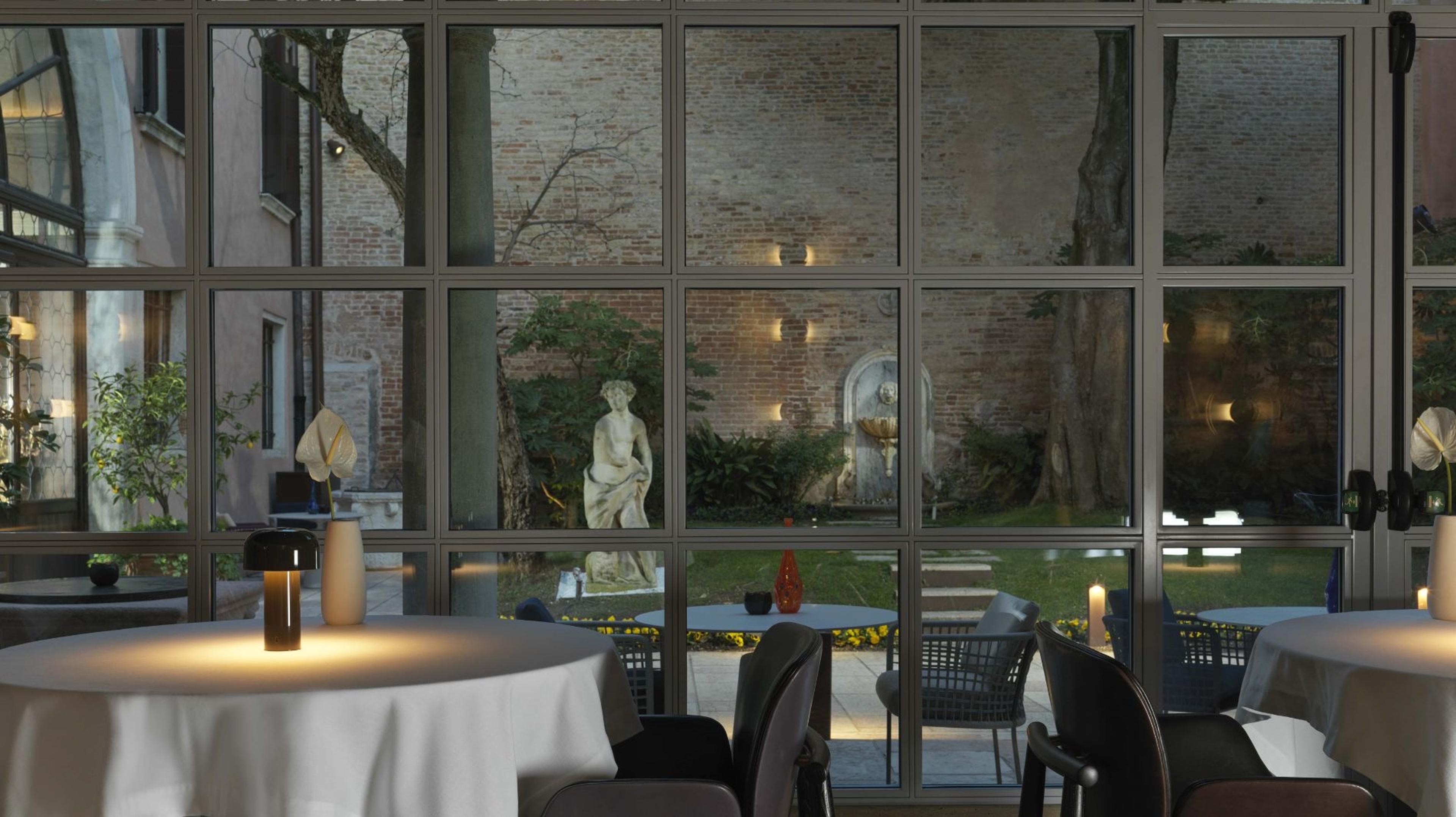 Tables set for dinner inside a restaurant, with glass doors looking out onto a courtyard garden with a statue, a wall fountain and string lighting at dusk.