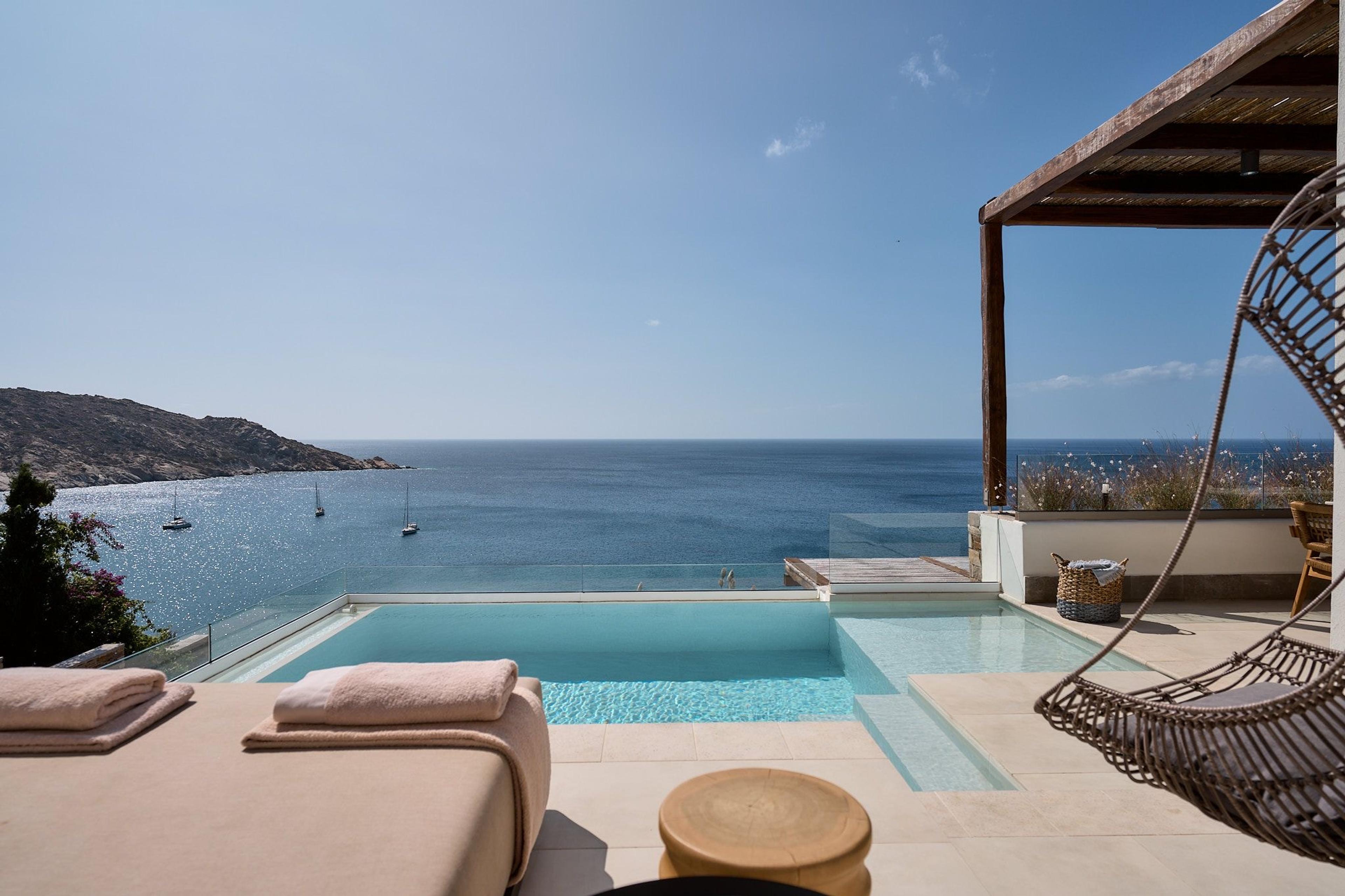 An infinity pool behind a glass rail, with sailing boats on the sea beyond, pink-towelled daybeds at the left, and a hanging wicker chair at the right.