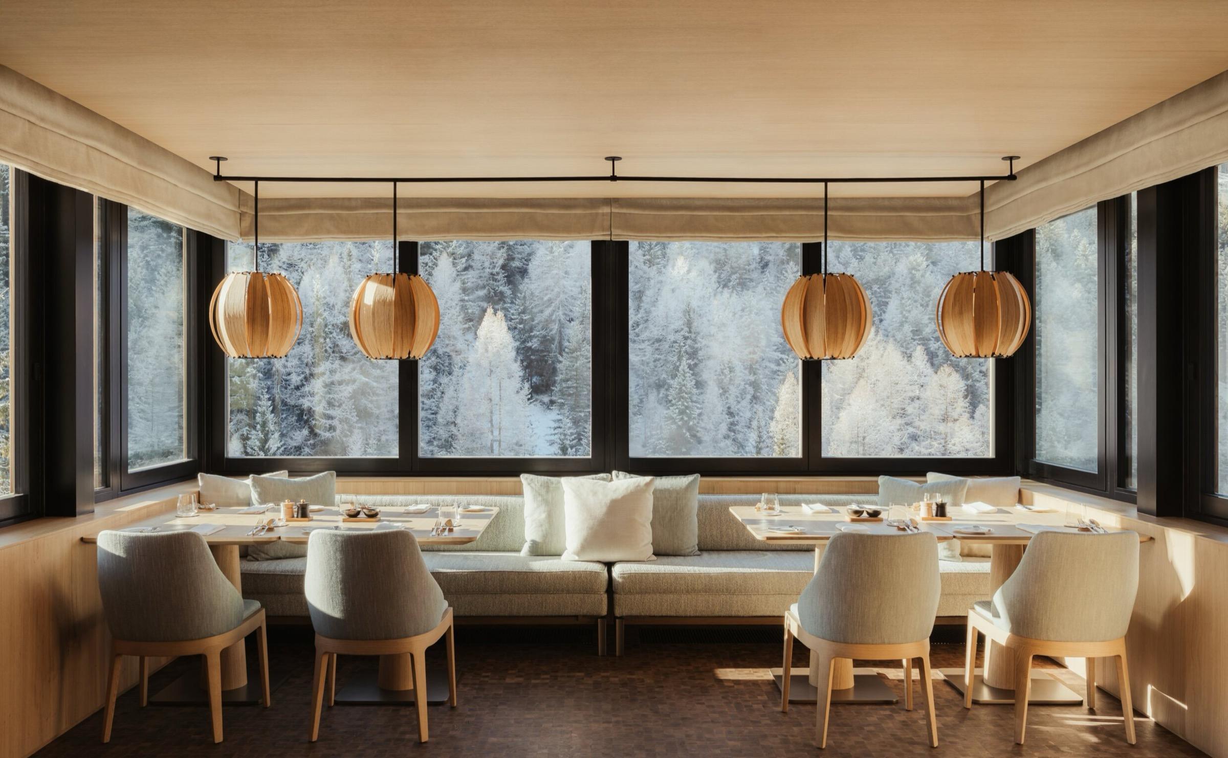 a dining room with round wooden pendant lights over banquette tables set with glasses and napkins, facing snowy pine trees through tall windows