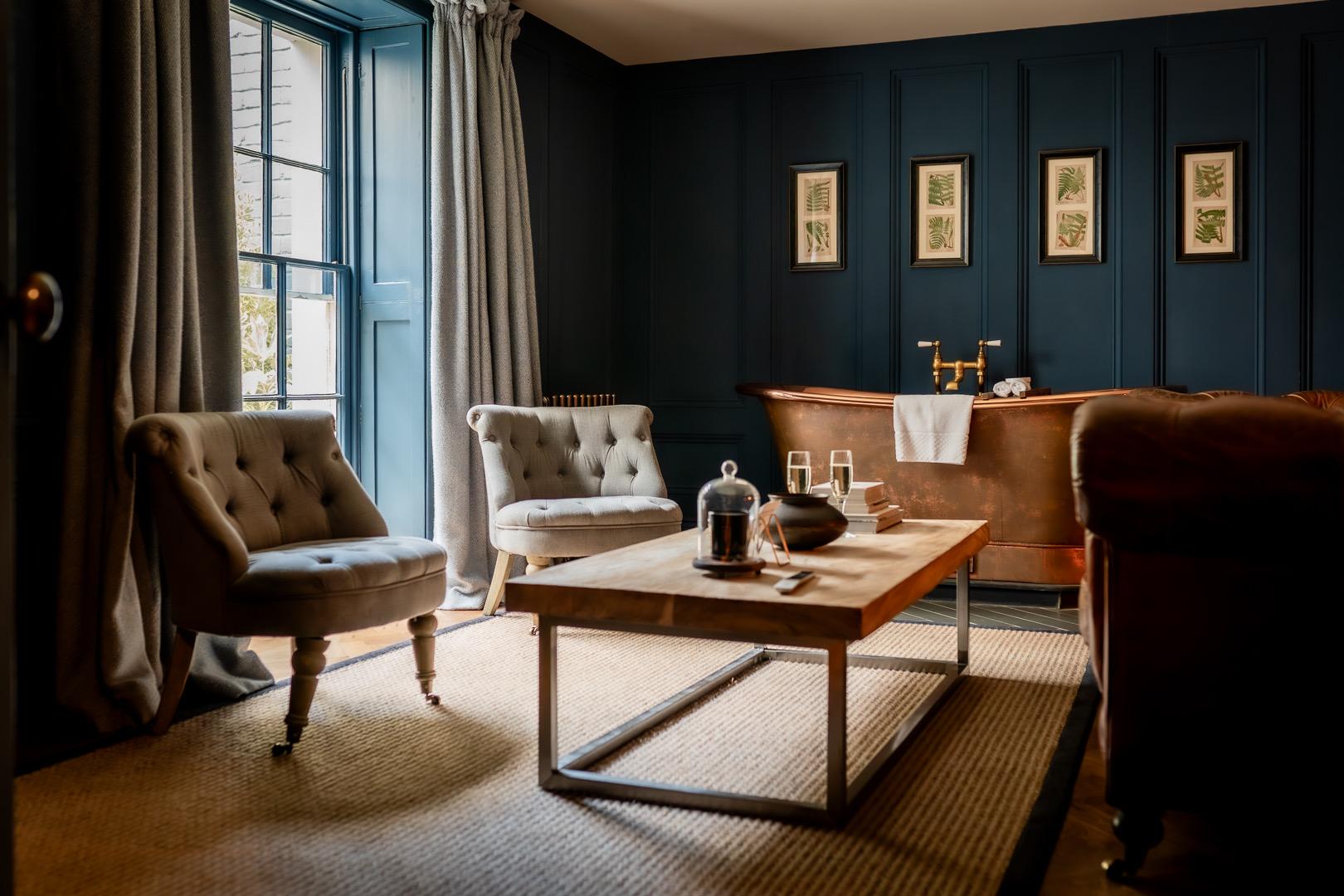 A sitting room with two tufted armchairs, a wooden coffee table and a copper freestanding bathtub against a dark blue panelled wall.