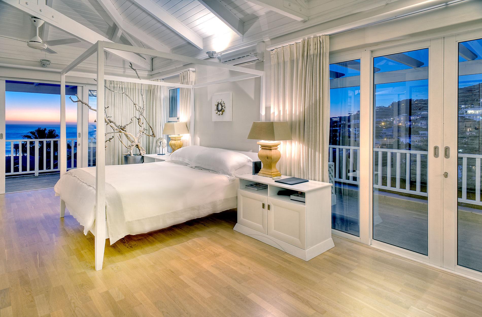 A white four-poster bed in a bedroom at dusk, with the sea and palm trees visible through the open glass doors to the balcony.