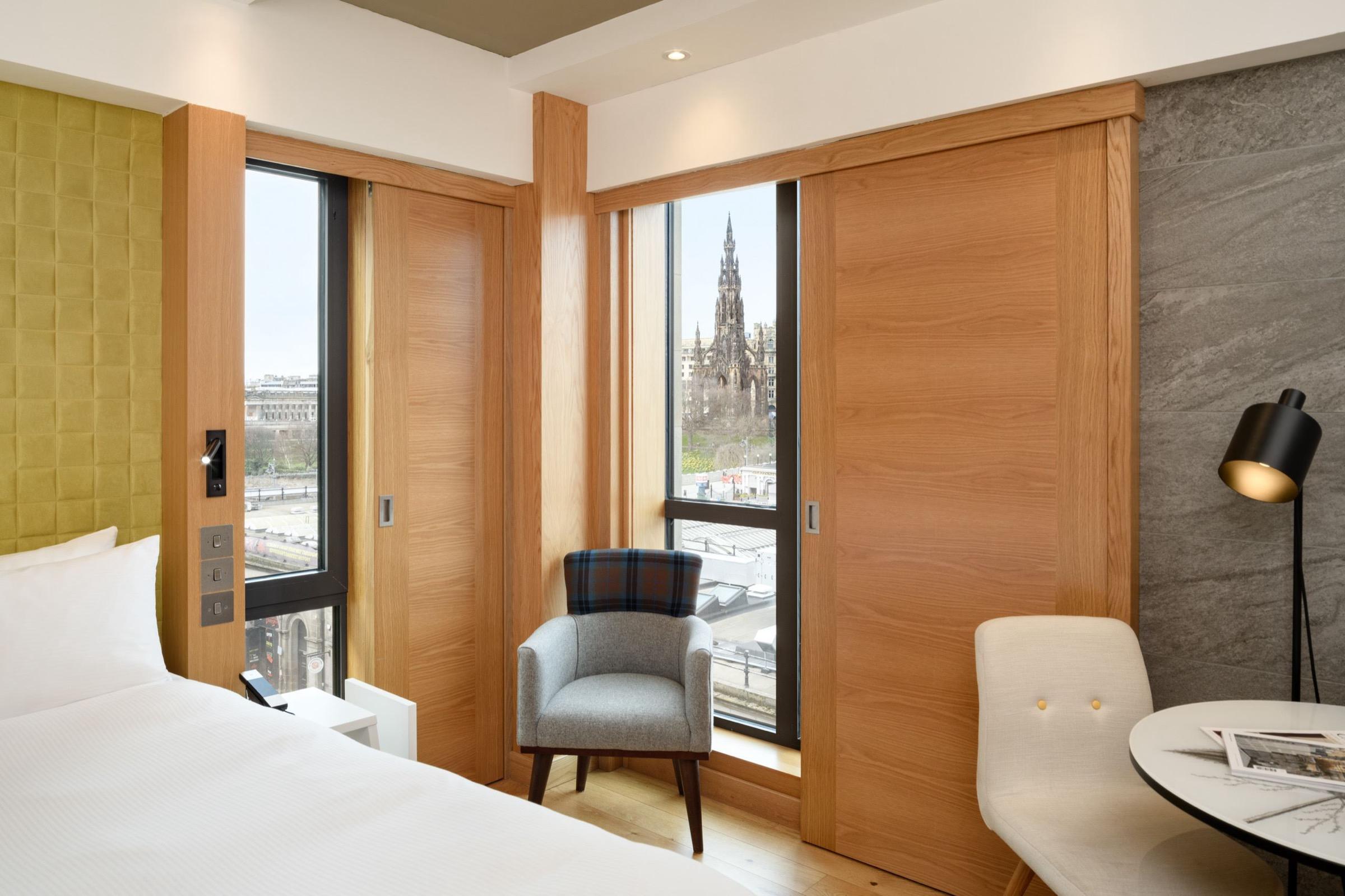 A bedroom with wooden sliding doors and a tartan armchair, with two windows looking out to the Scott Monument and Princes Street Gardens.